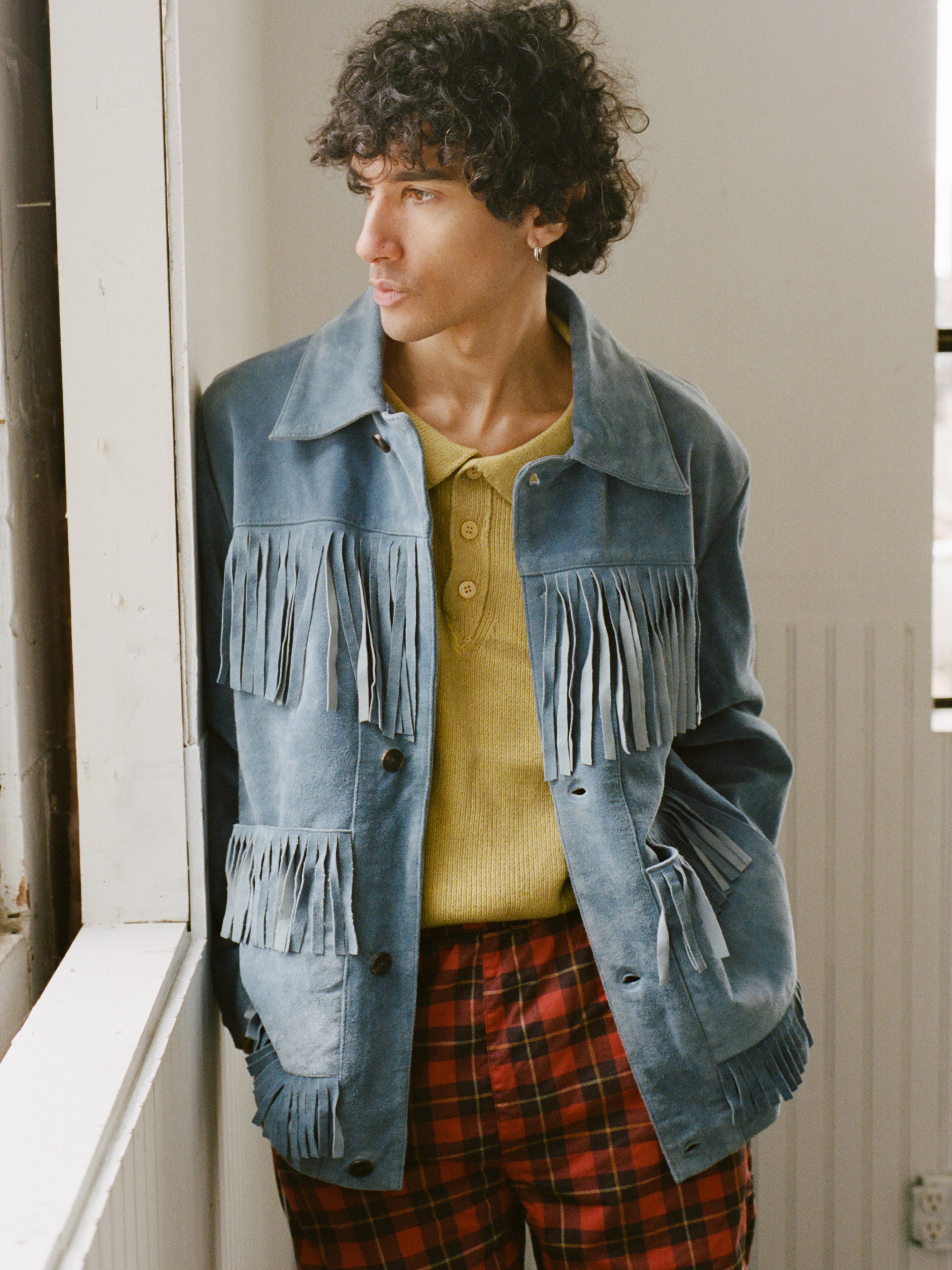 A person with curly hair wears FOUND's Western Suede Fringe Jacket in indigo blue, paired with a yellow shirt and red plaid pants, standing by a window and looking to the side.