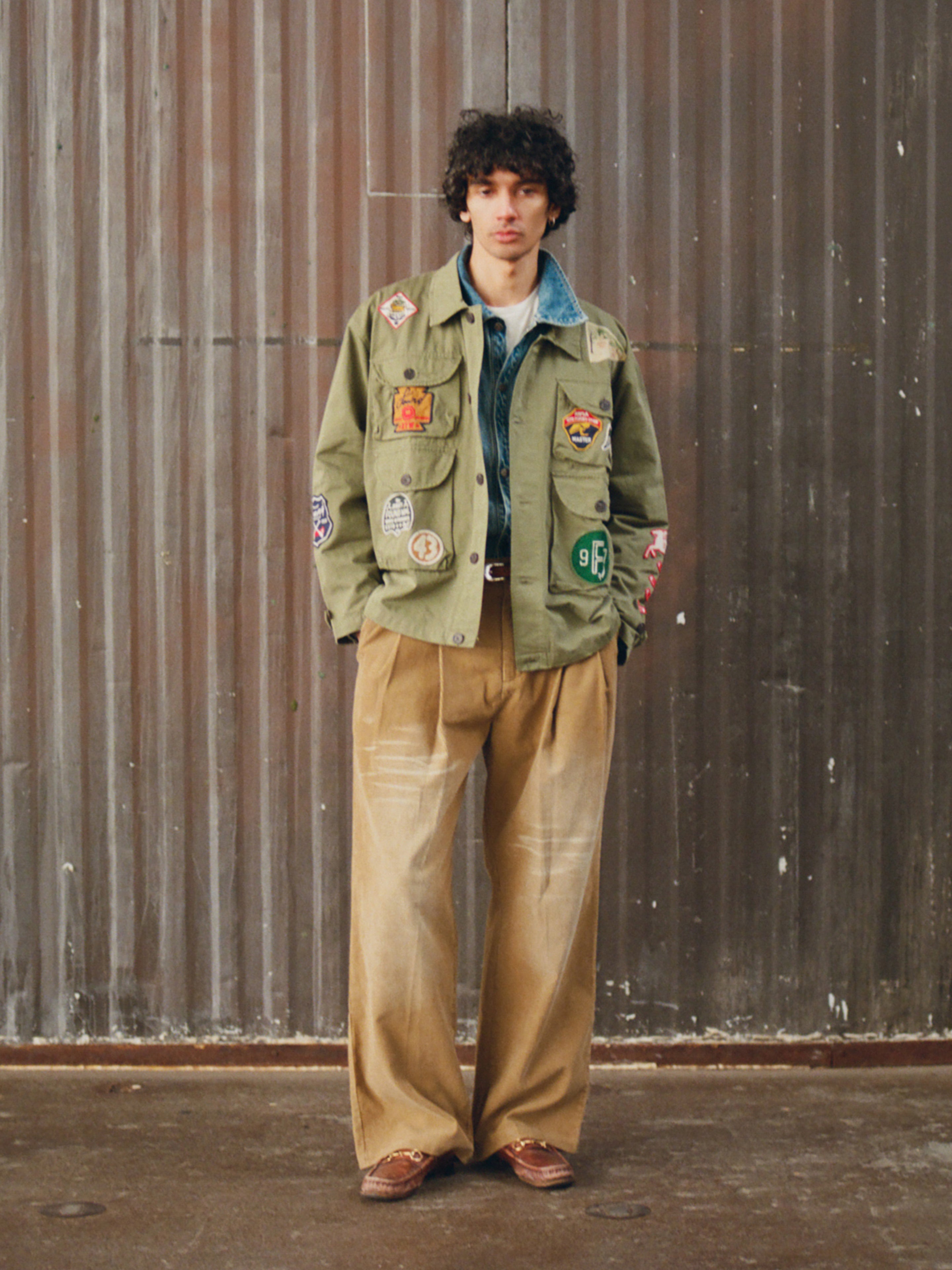 A person stands indoors against a corrugated metal wall wearing the FOUND Herringbone Utility Patchwork Jacket in olive green, paired with a denim shirt, wide tan pants, and brown shoes.
