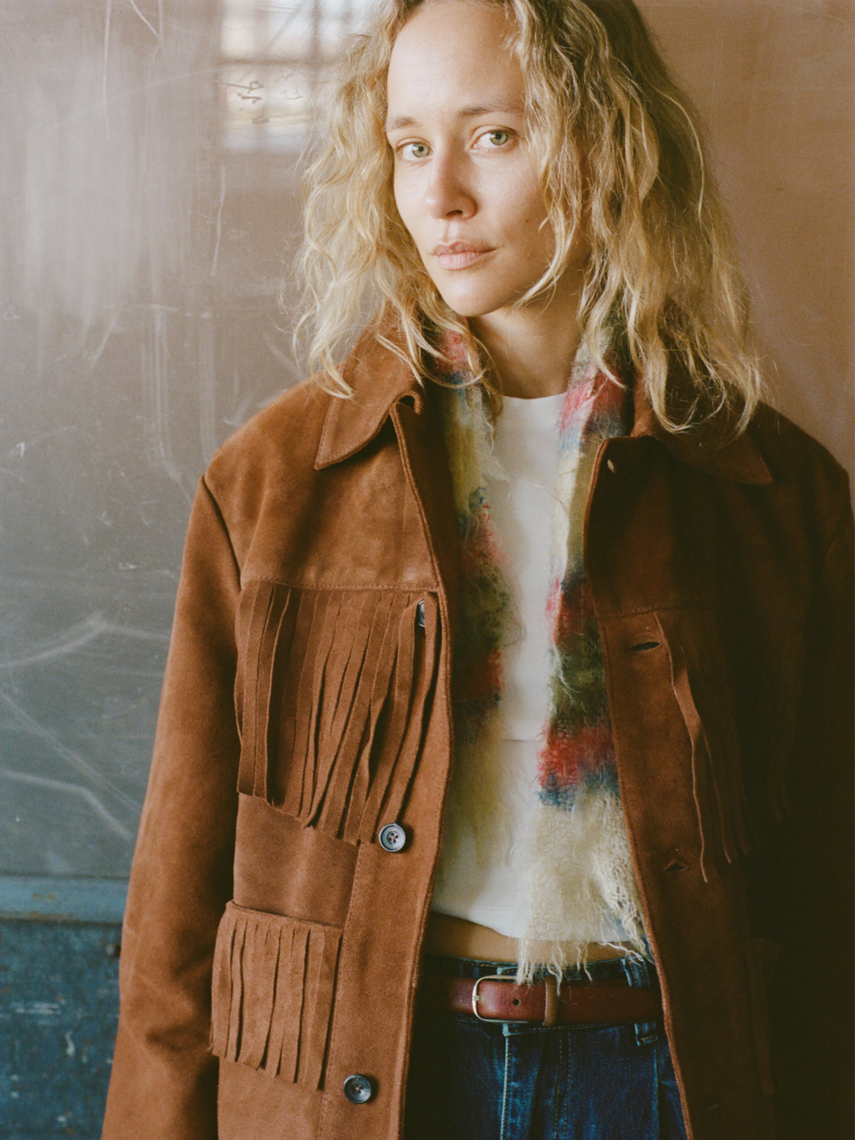 A woman with wavy blonde hair wears the FOUND Western Suede Fringe Jacket over a white top and blue jeans, accessorized with a scarf, standing against a plain background.