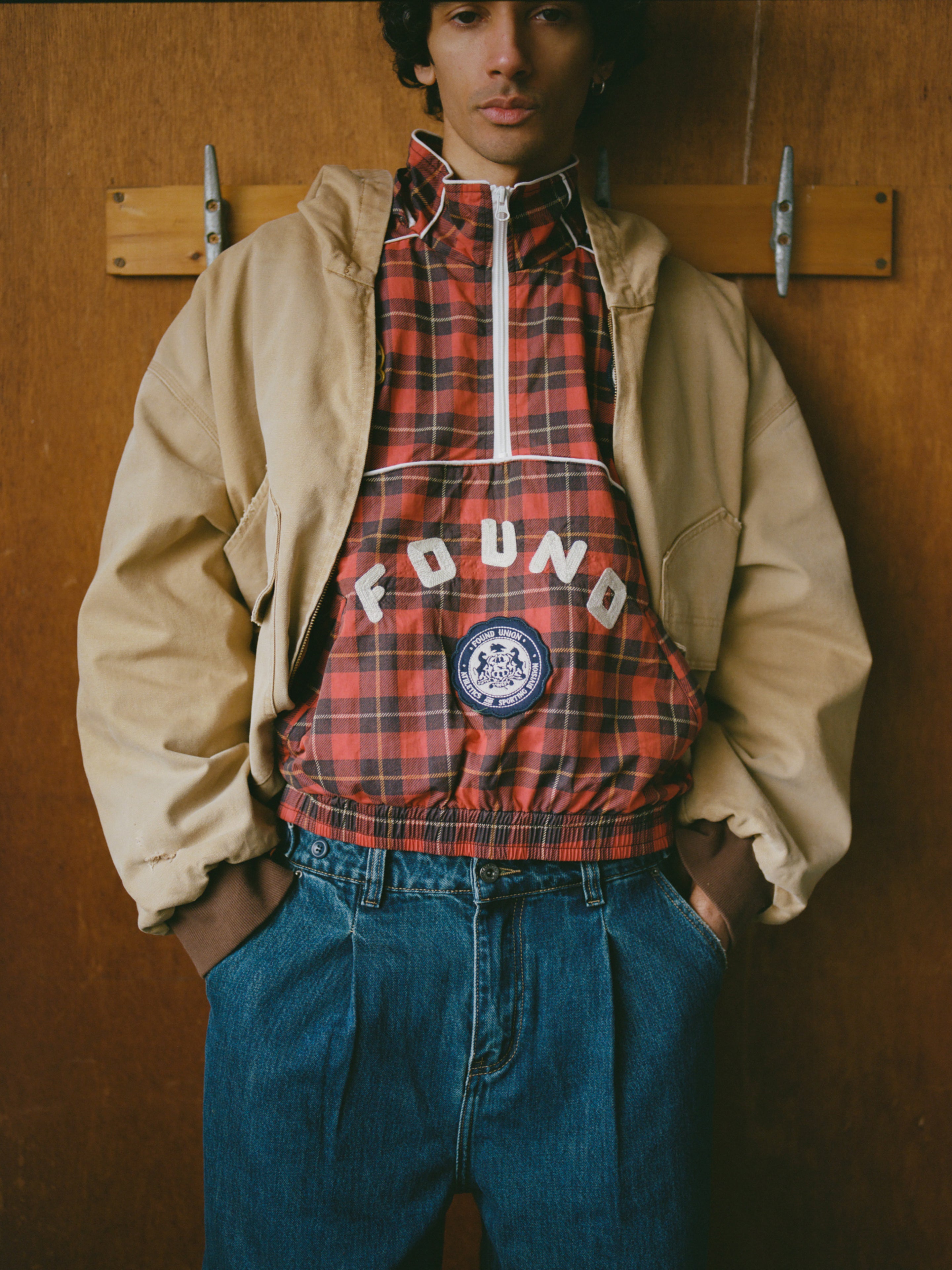 A person wearing FOUND's Red Plaid Track Jacket, featuring the brand's name and a circular emblem, stands against a wooden wall with hands in blue jeans pockets.
