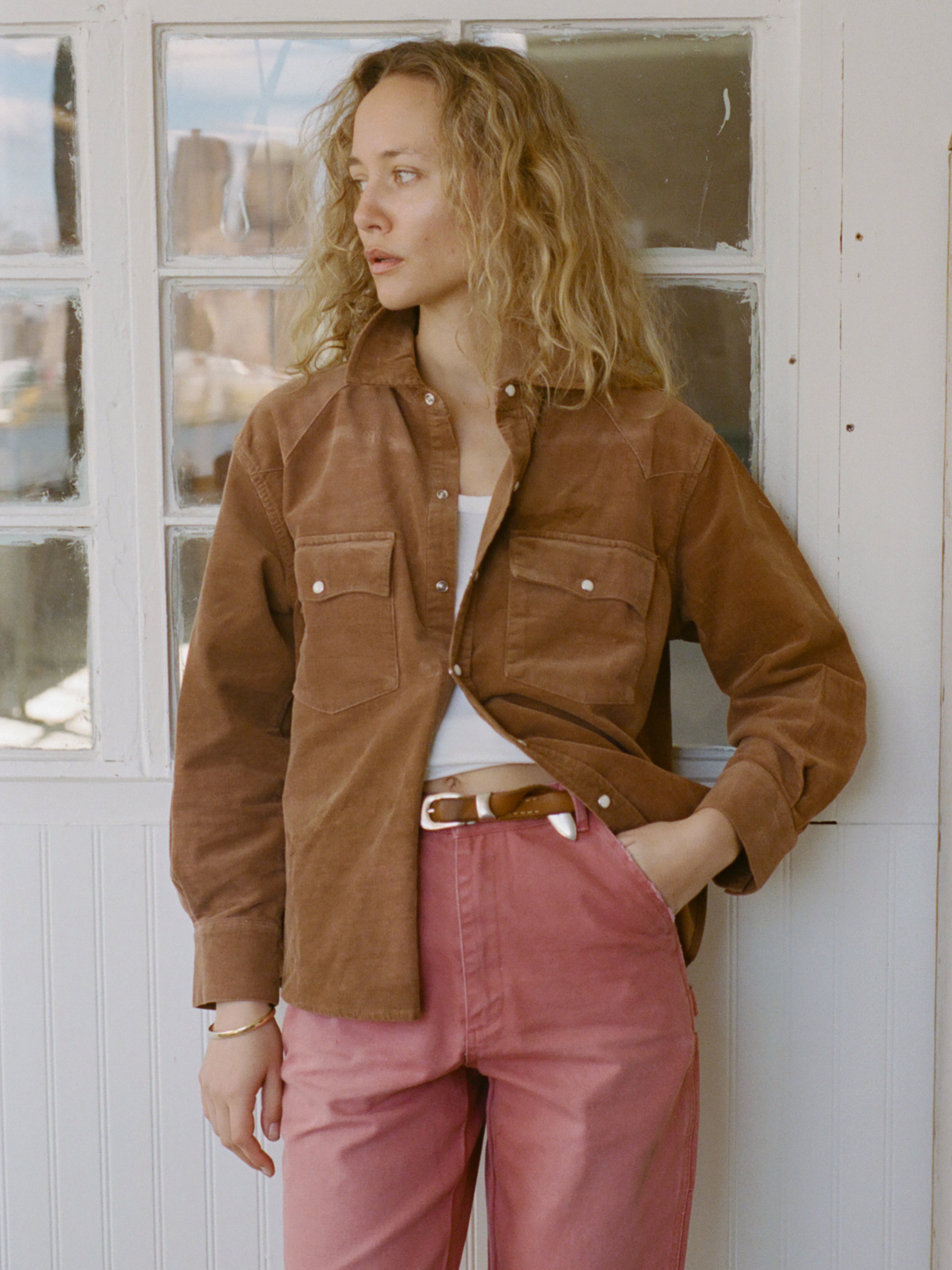 A woman with curly blonde hair stands indoors, wearing the FOUND Western Corduroy Shirt over a white top and pink pants, one hand in her pocket.