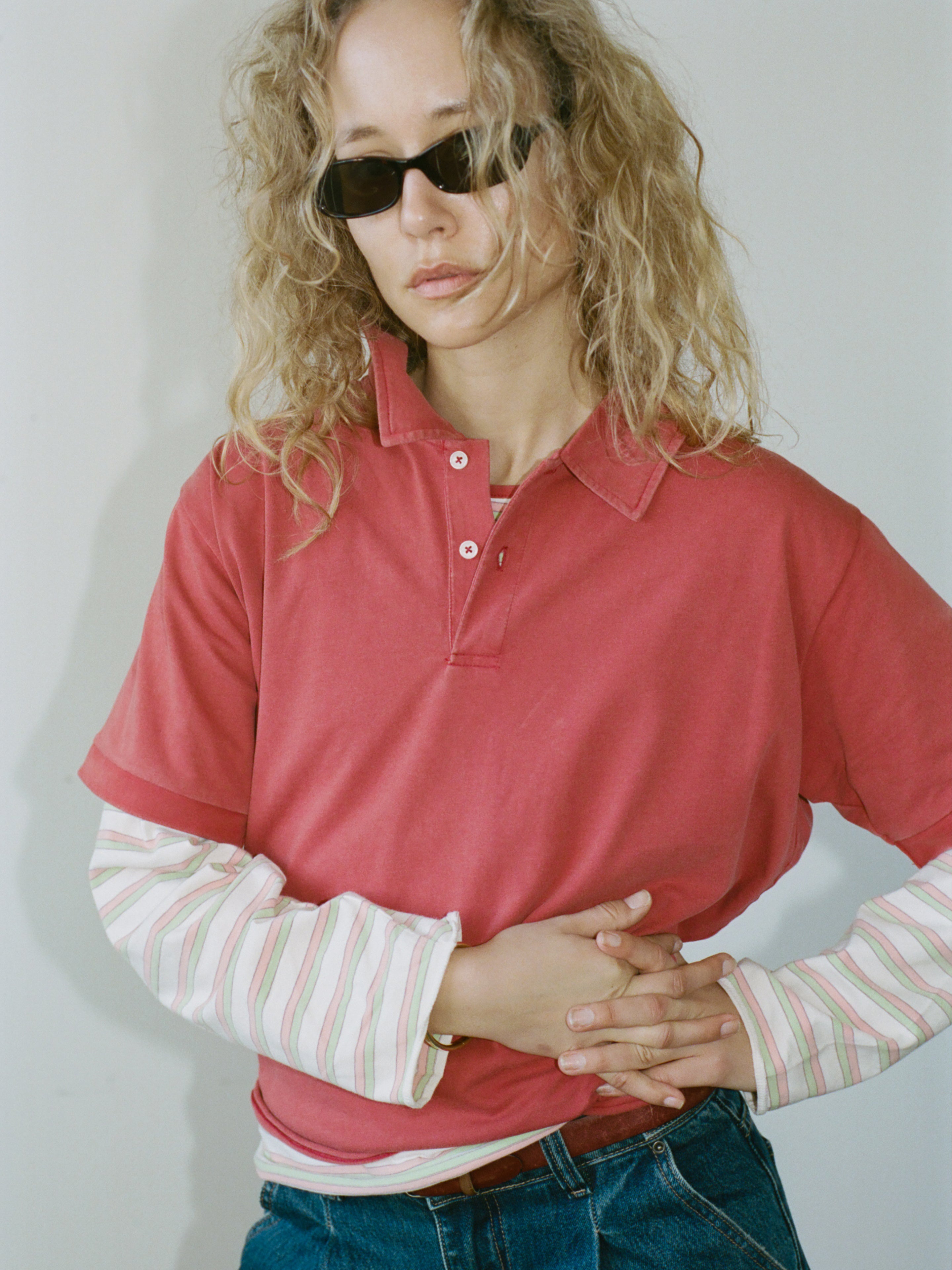 A person with curly blond hair and sunglasses wears the FOUND Double Layer Stripe Polo over blue jeans, standing against a plain background.