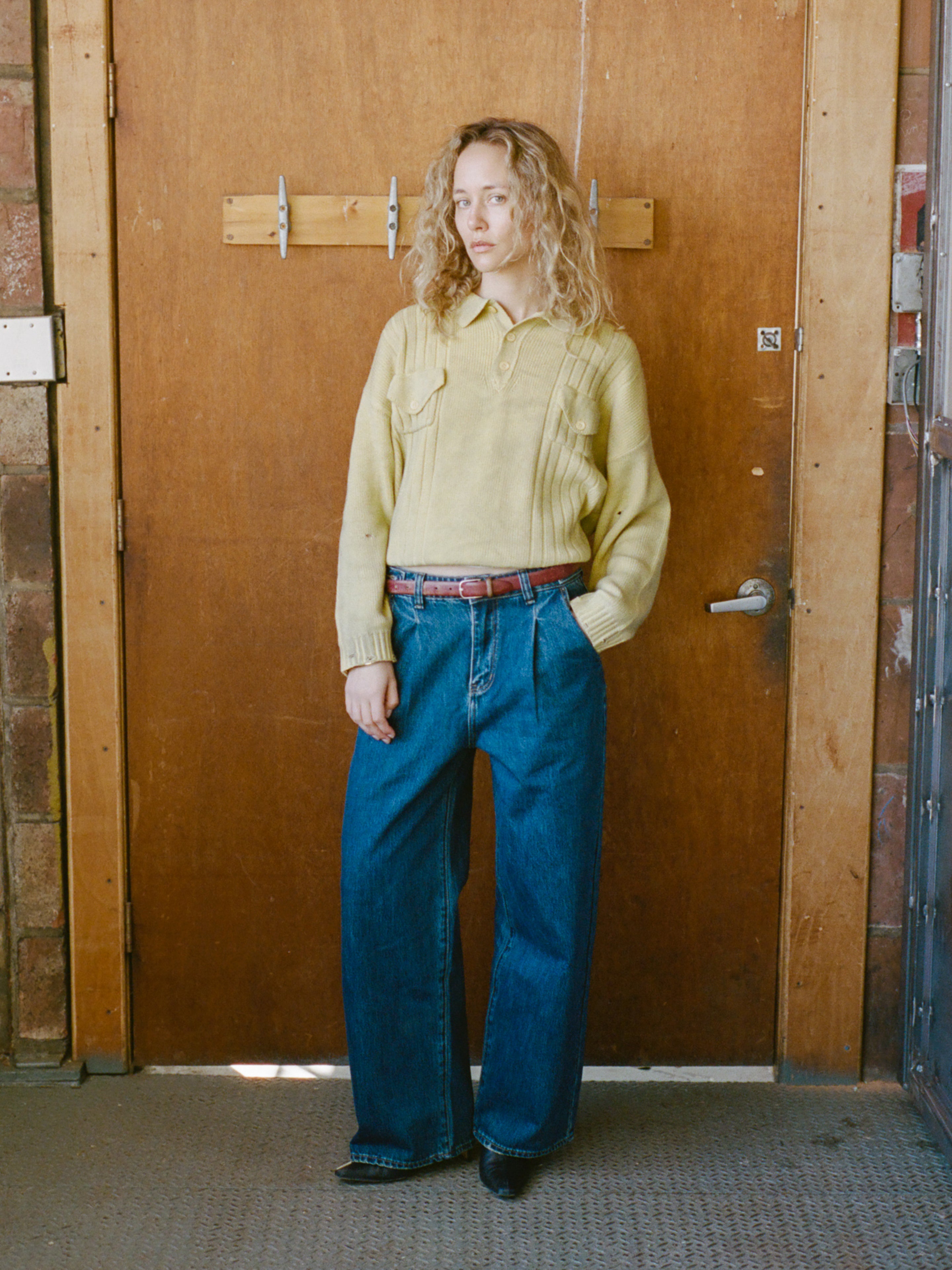 A person with curly hair stands indoors by a wooden door, wearing the FOUND Distressed Pullover Knit in yellow, blue wide-leg jeans, and a brown belt.