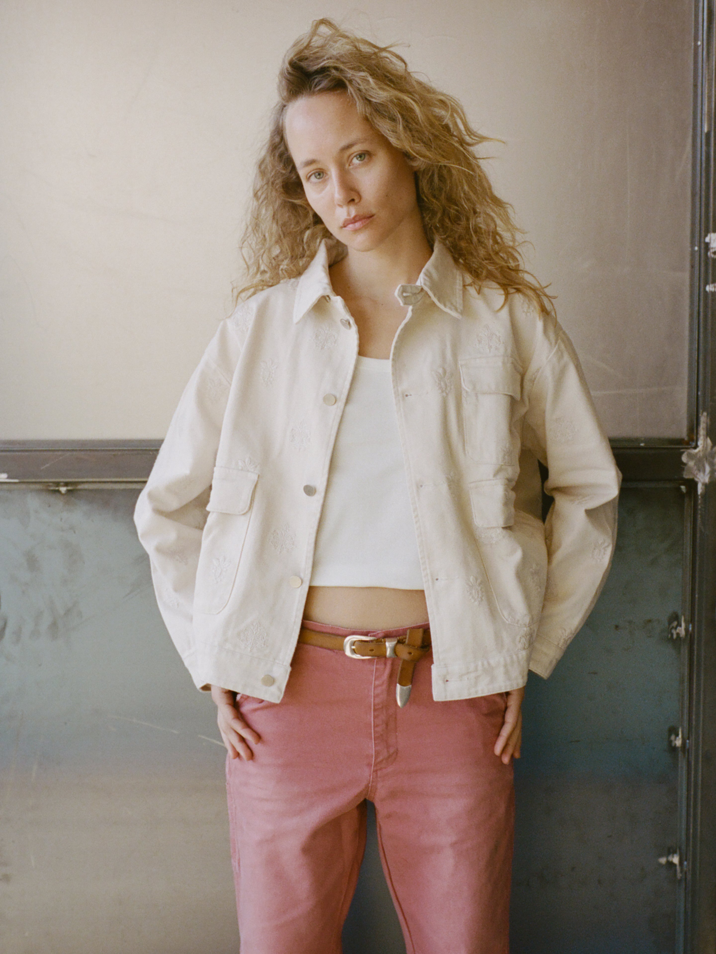 Woman with wavy hair wearing the Found Embroidered Motif Jacket over a white top and pink pants, standing against a metallic background with hands in her pockets.
