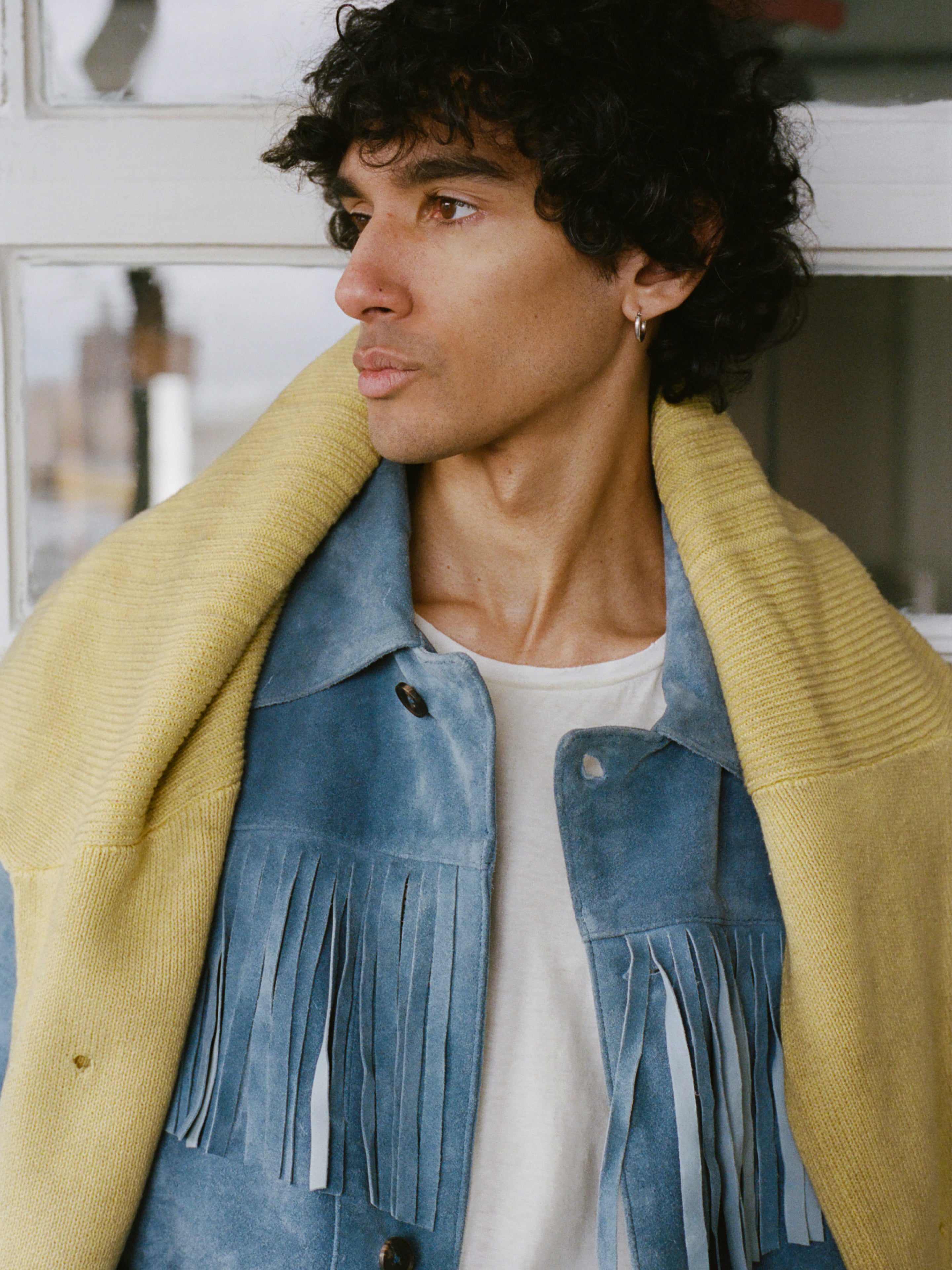 A person with curly hair wearing the FOUND Western Suede Fringe Jacket in indigo blue, a white shirt, and a yellow knit draped over their shoulders looks to the left.