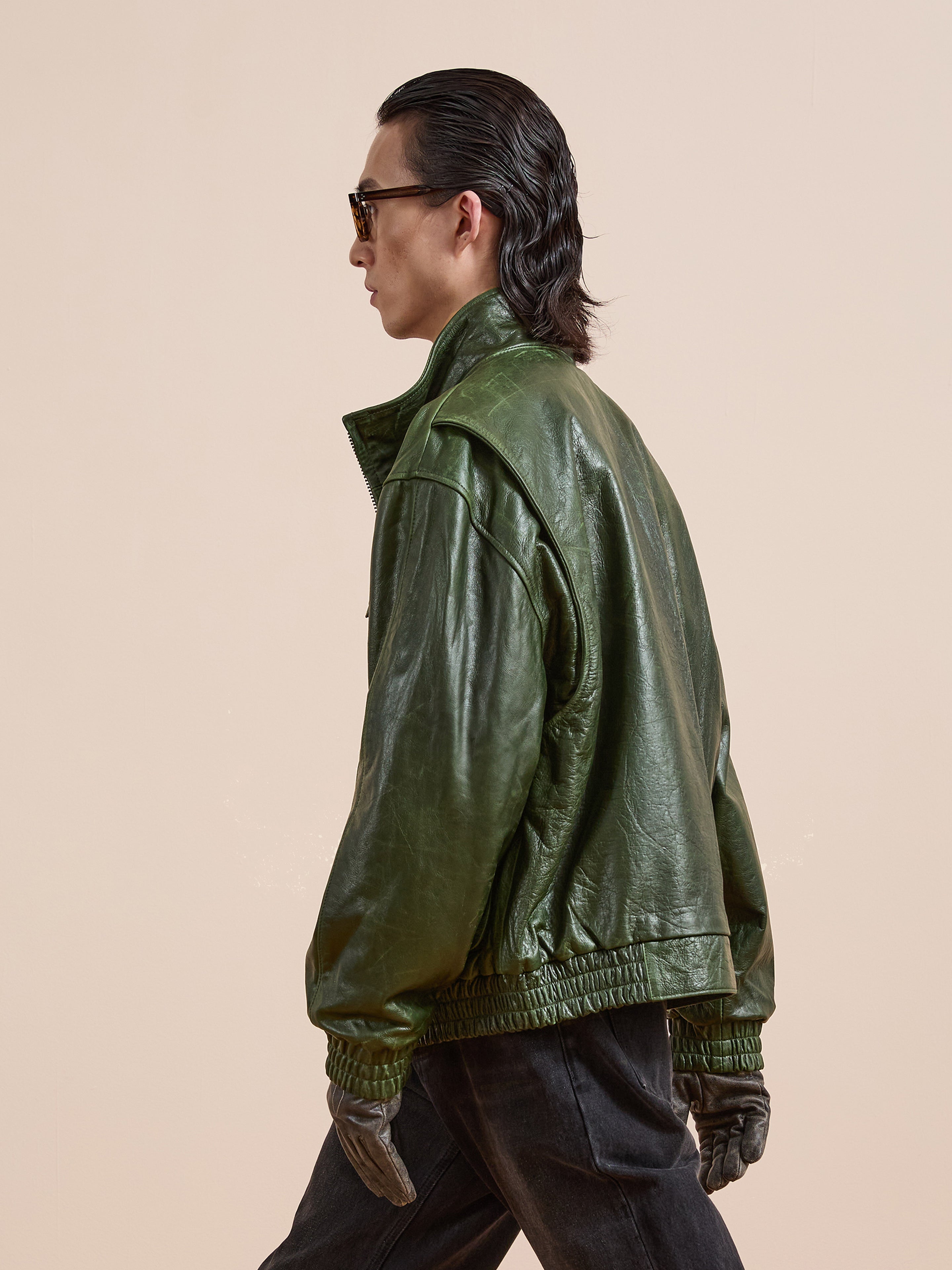 A person wearing the FOUND Forest Leather Bomber Jacket, paired with sunglasses and dark gloves, walks to the left against a plain beige background.