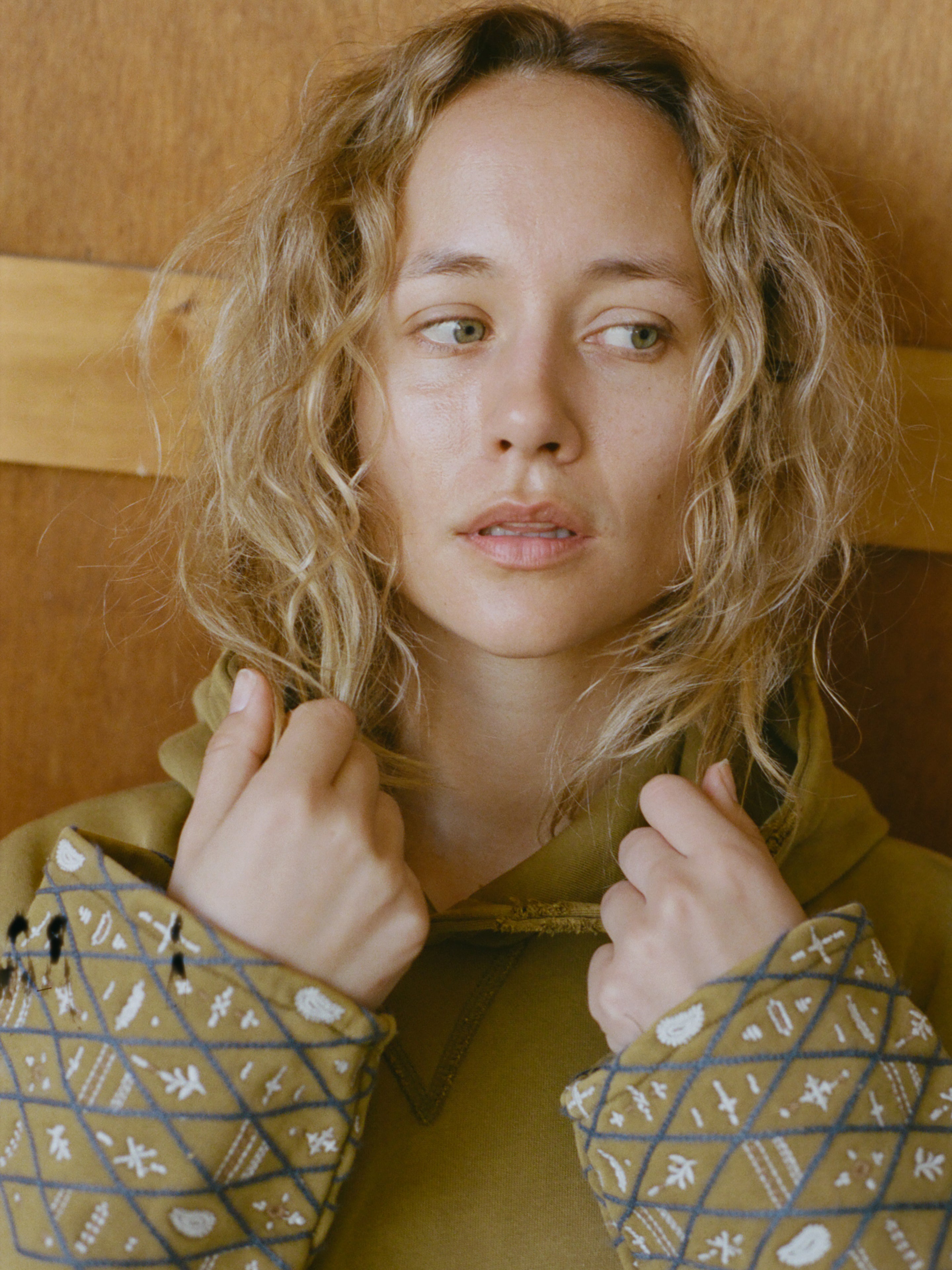 A person with wavy blond hair looks to the side, holding the collar of a FOUND Embroidered Border Hoodie in both hands, standing against a wooden backdrop.