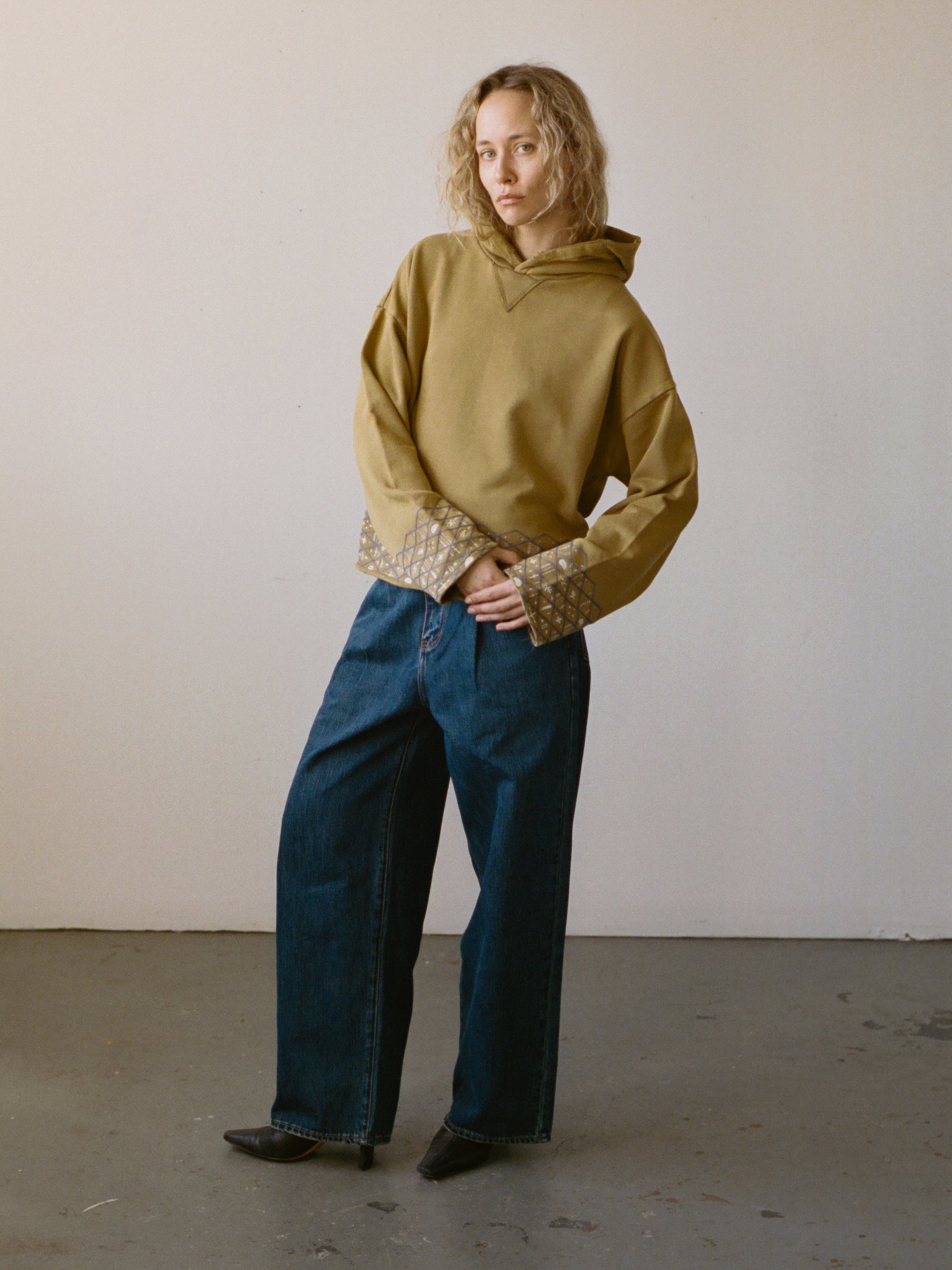 A person stands indoors wearing the FOUND Embroidered Border Hoodie, featuring detailed cuffs, paired with wide-leg blue jeans and black pointed shoes against a plain light-colored wall.