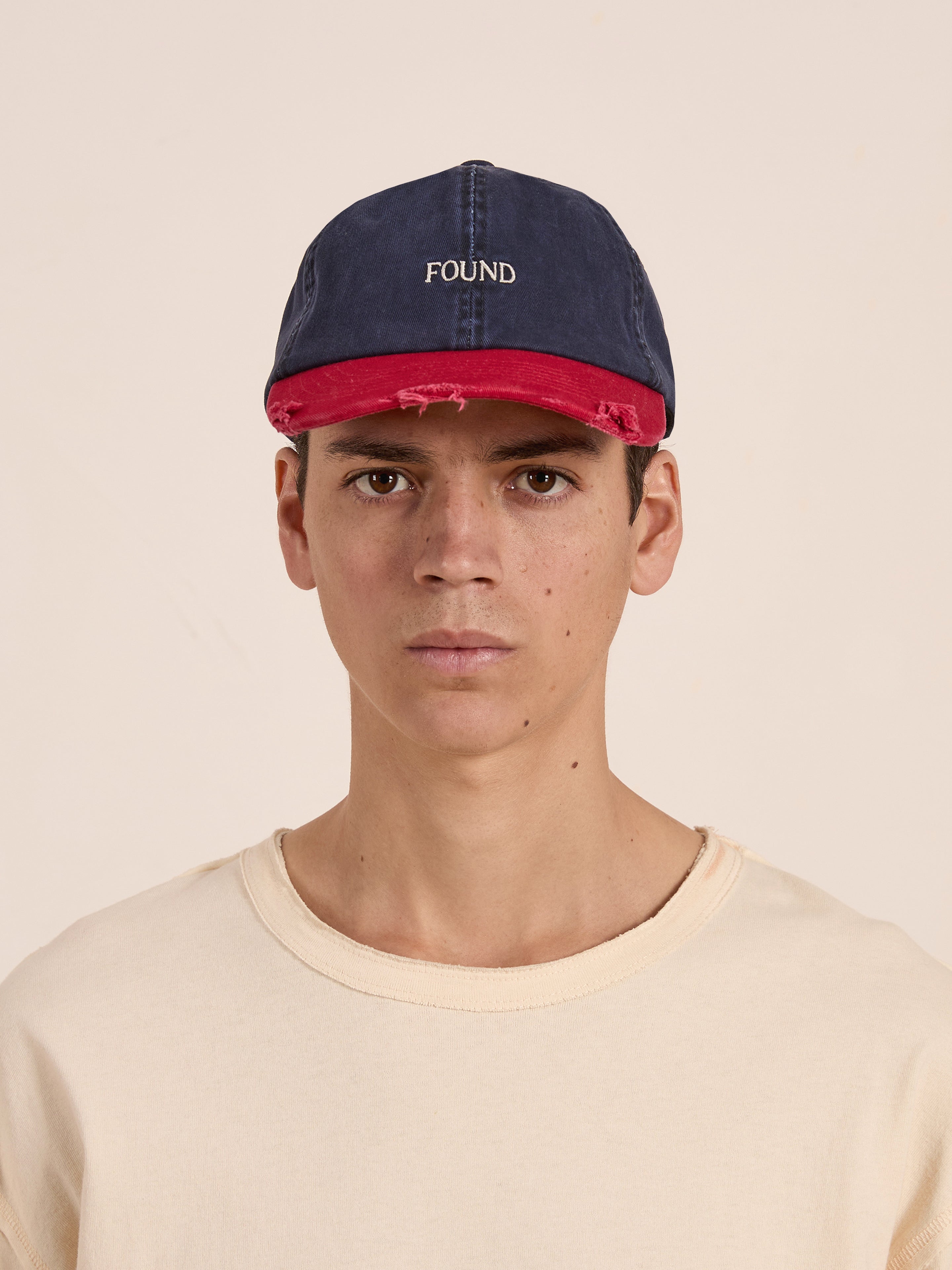 Someone in a beige shirt wears a Two-Tone Logo Cap from FOUND, featuring the brand's logo prominently on the red brim. The cap's two-tone design adds contrast to the neutral outfit while maintaining a composed look.