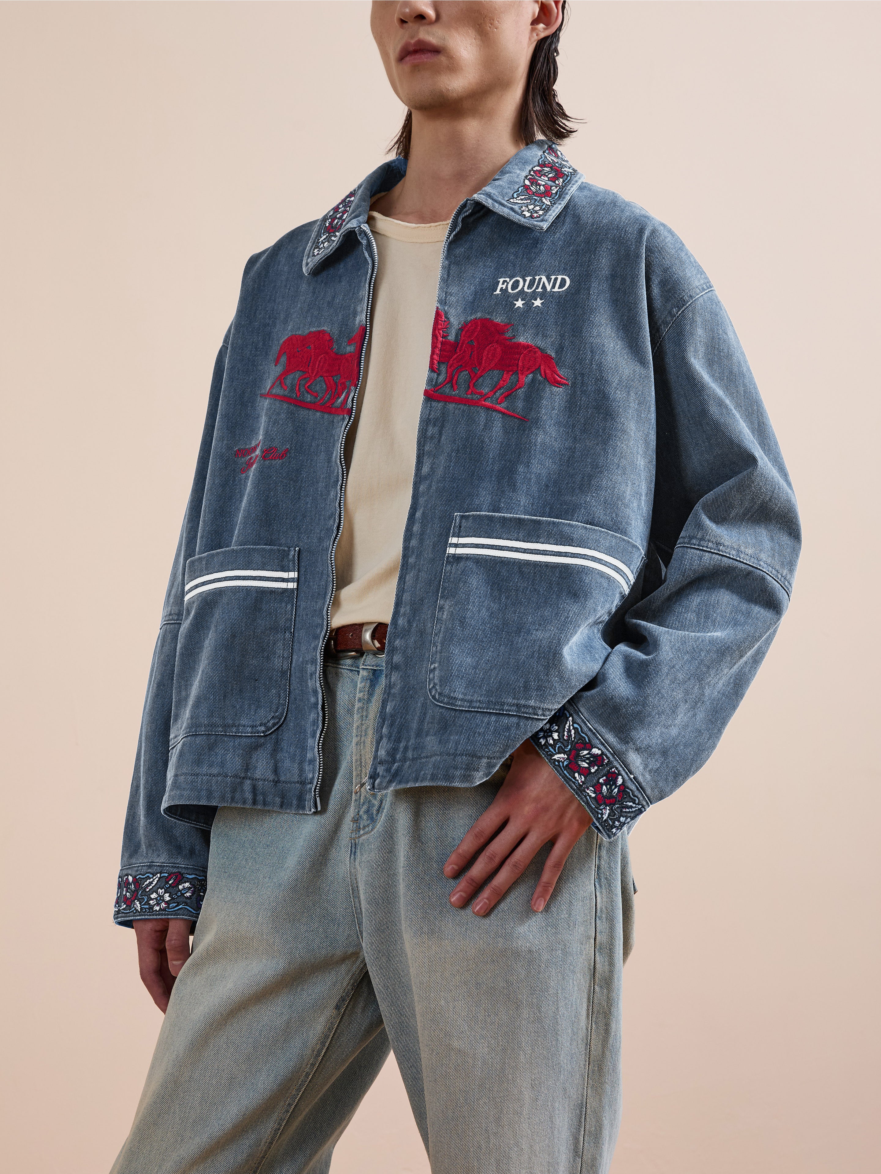 A person in the Found Jockey Club Embroidered Jacket—blue with racing horse embroidery, patterned collar and cuffs—and matching relaxed-fit blue jeans, stands against a neutral background.
