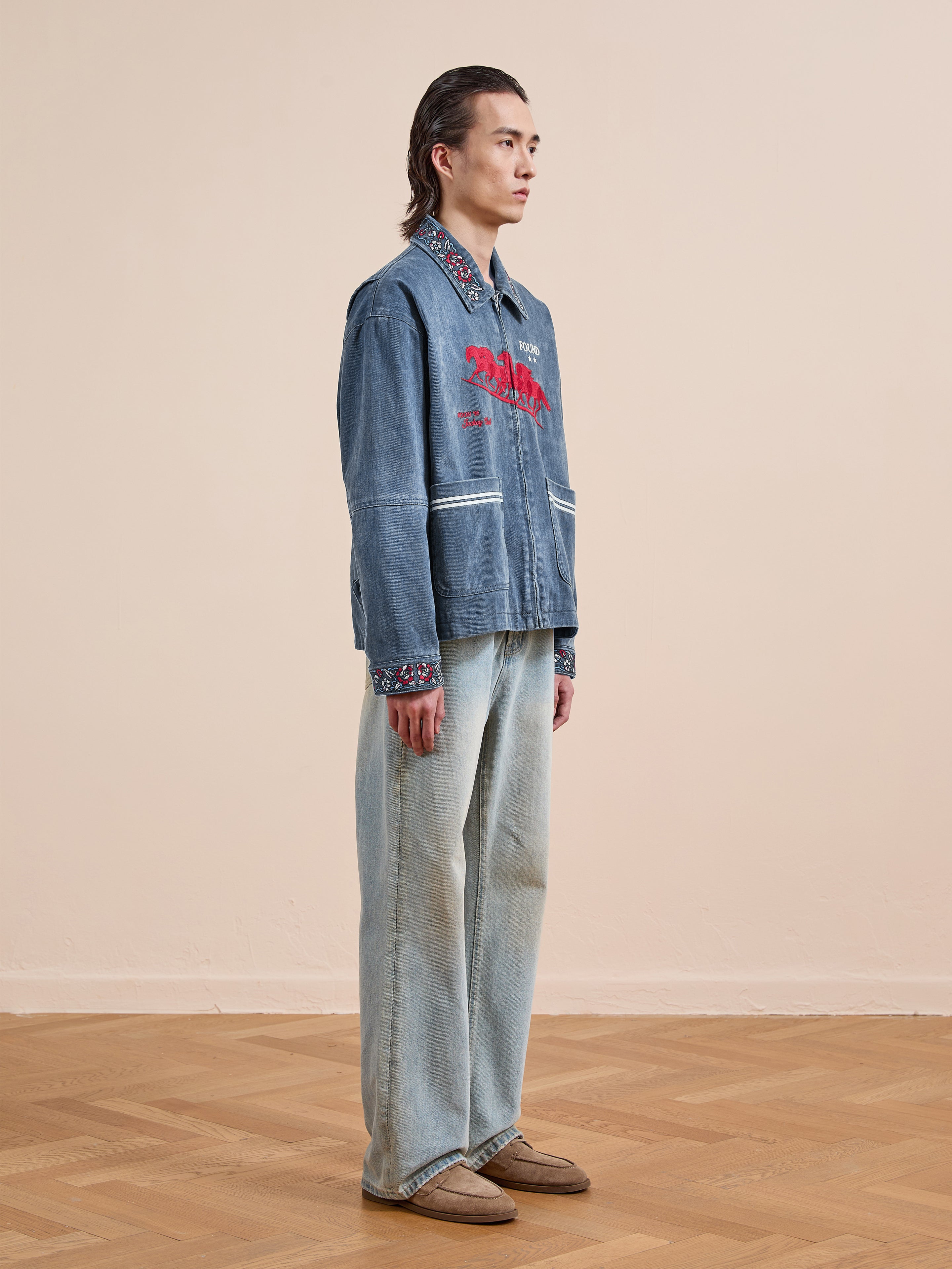 A person stands sideways on a wooden floor, wearing the Found Jockey Club Embroidered Jacket, light blue wide-leg jeans, and brown shoes, against a plain beige wall.