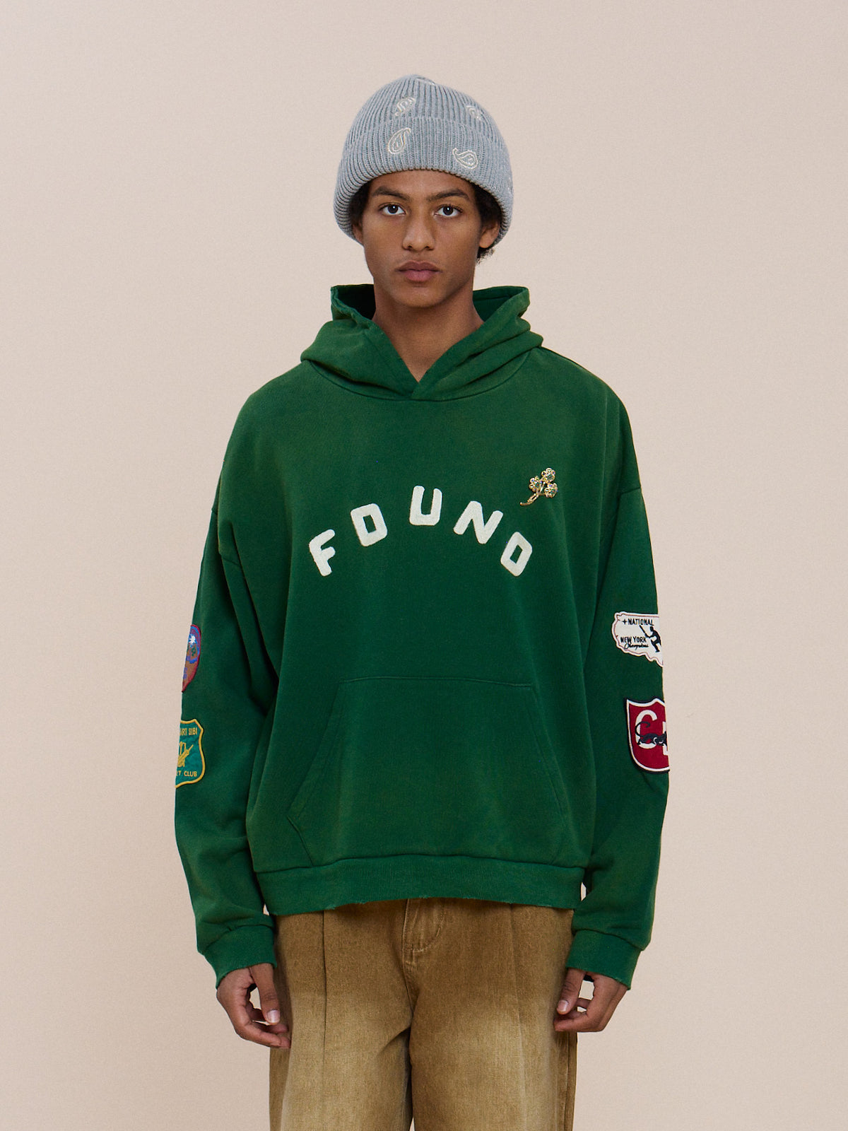 Person wearing a gray knit beanie and FOUND Found Logo Patch Hoodie in vintage forest green, paired with tan pants against a plain light background.