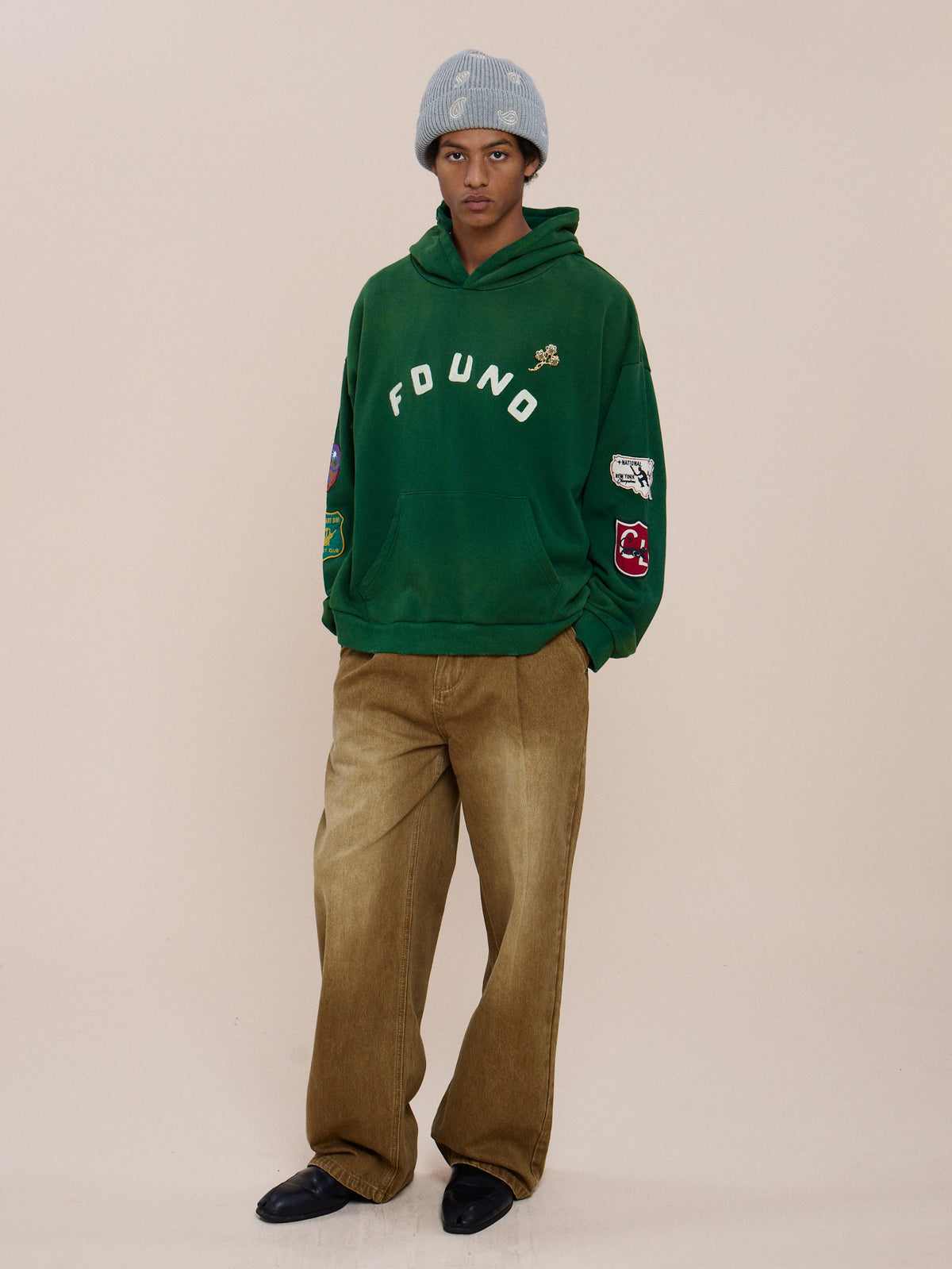 Person wears a vintage forest green Found Logo Patch Hoodie by FOUND, unisex relaxed fit brown pants, black shoes, and a gray beanie against a plain beige background.