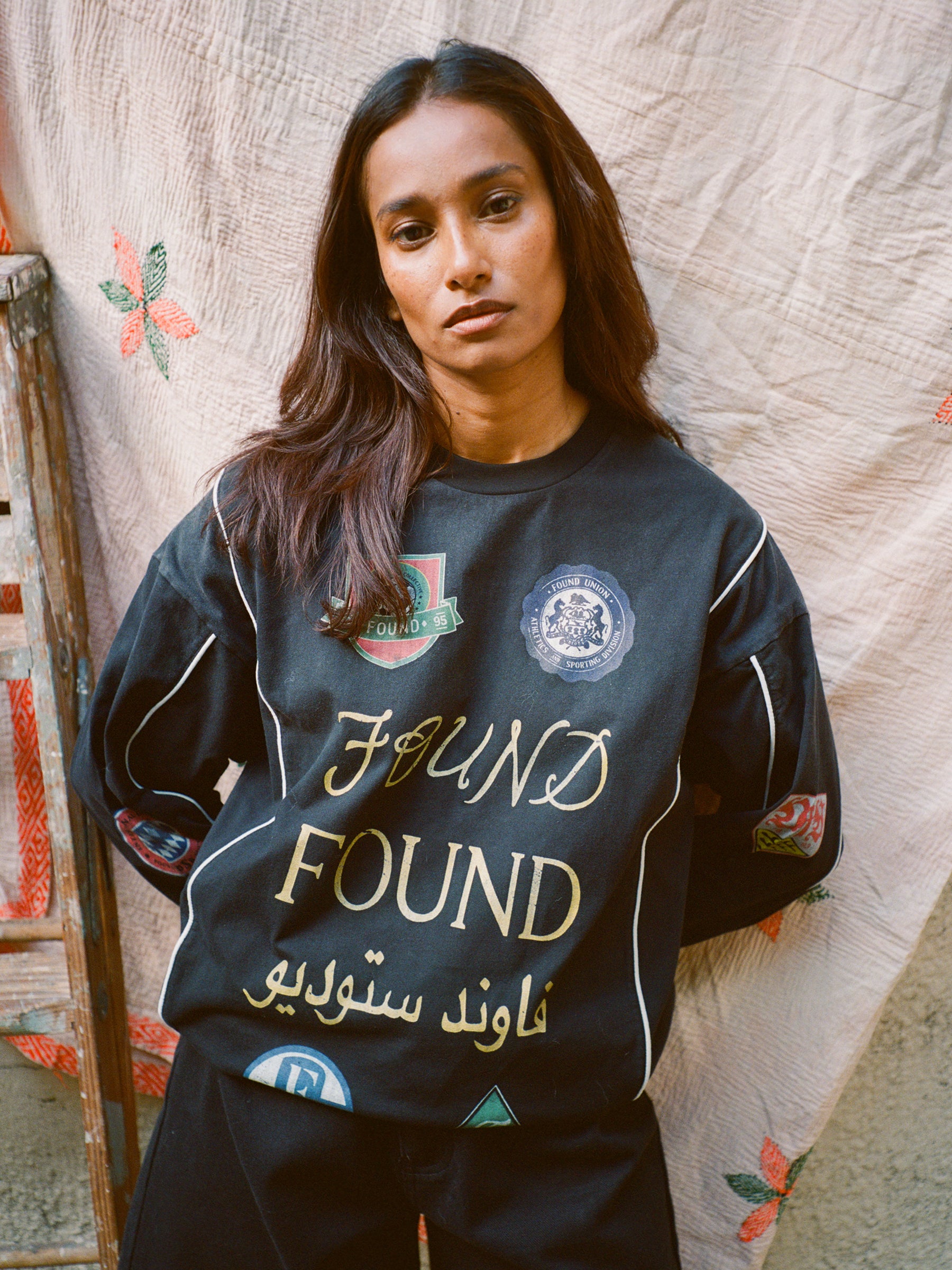 A confident person, donning the FOUND Multi Language LS Jersey with stylish patches, stands against a patterned backdrop. The retro outfit exudes timeless style, enhanced by a wooden ladder beside them.