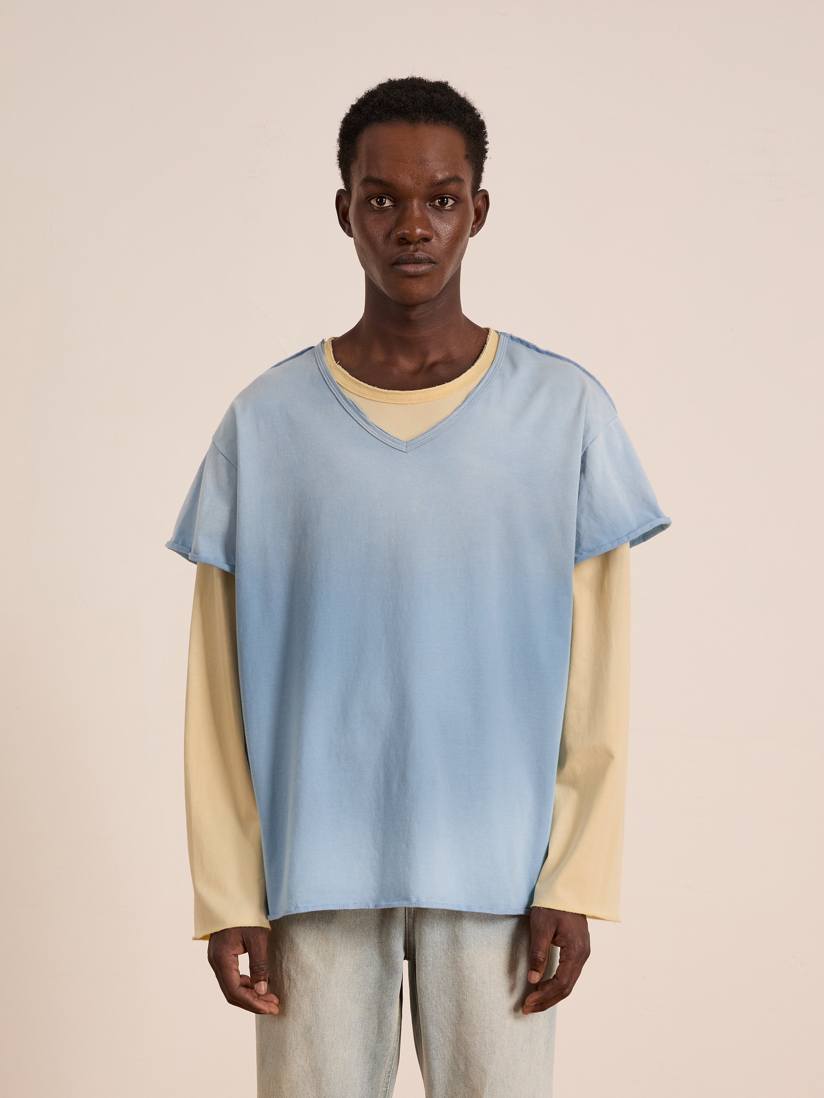 A person stands against a plain background, wearing FOUND’s Double Layer LS Tee—a light blue short sleeve over beige unisex long sleeve—paired with light-colored pants.