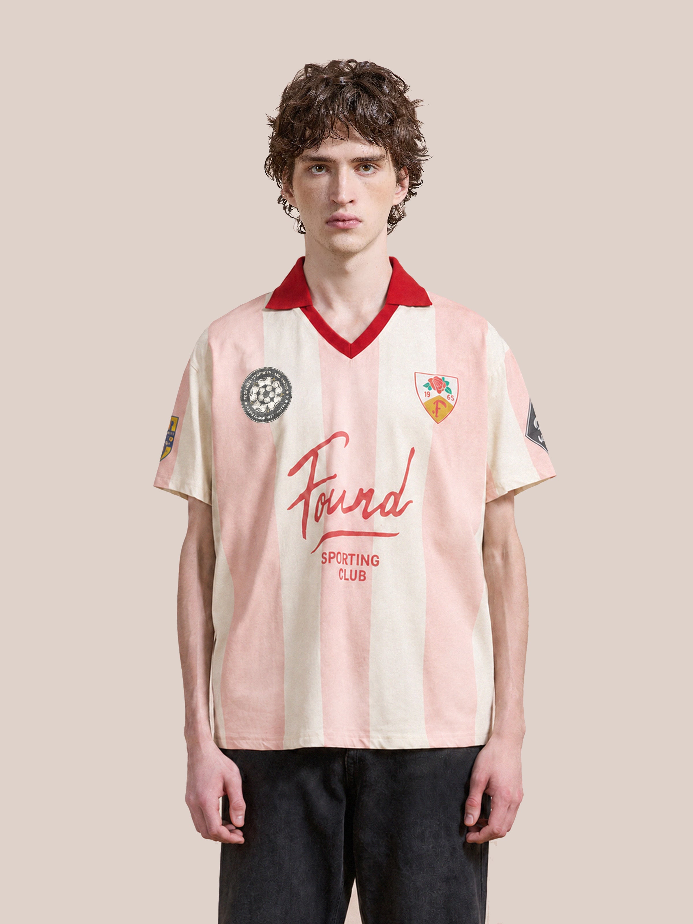 A young person with curly hair models the FOUND Found Club Short Sleeve Jersey—a unisex relaxed fit top featuring light pink and white stripes, a red collar, assorted patches, and the “Found Sporting Club” logo—while standing against a plain background.
