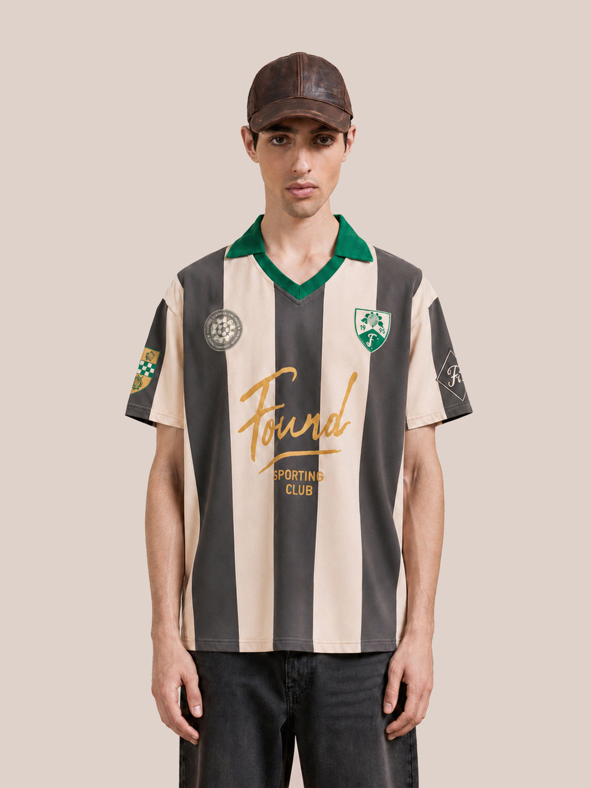 A person wearing the FOUND Found Club Short Sleeve Jersey—a black and beige striped vintage football shirt with a green collar—paired with a brown cap and black pants, stands facing forward against a plain background. This unisex, relaxed fit jersey from FOUND adds a classic touch to their look.