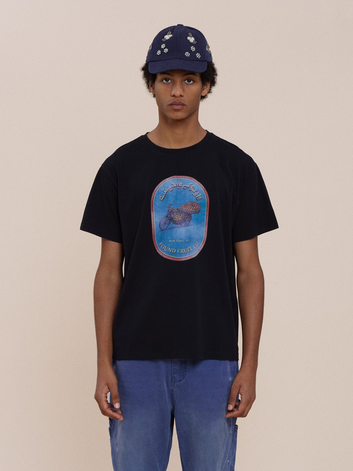 Wearing a navy cap, blue jeans, and the FOUND Fruit Co Tee, a unisex relaxed fit vintage fruit stand shirt, they stand against a plain beige background.