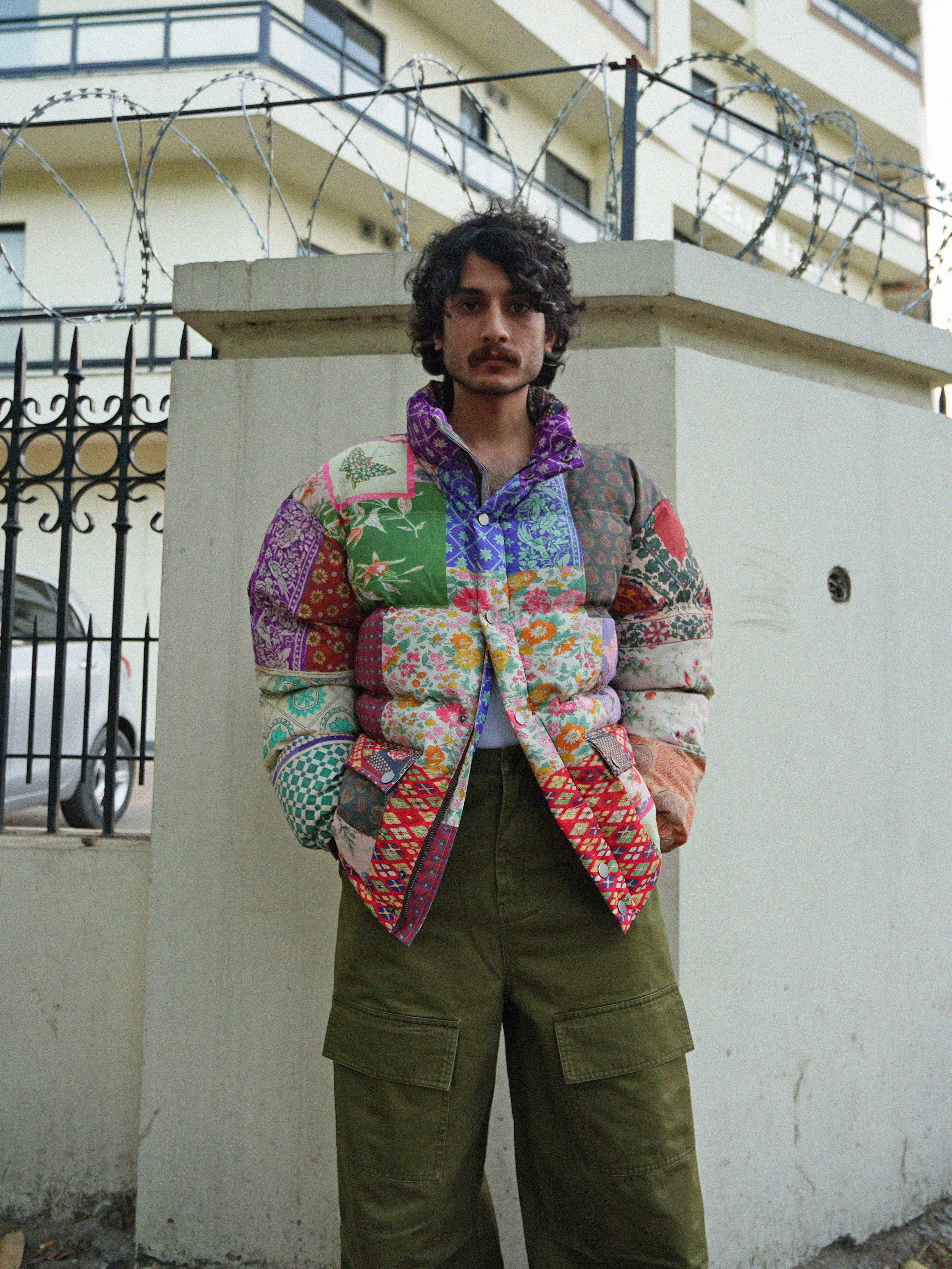 A person wearing the Gardenia Tapestry Puffer Jacket by Found, with a design reminiscent of tapestries and quilts, and olive pants stands in front of a white wall topped with barbed wire, gazing directly at the camera.