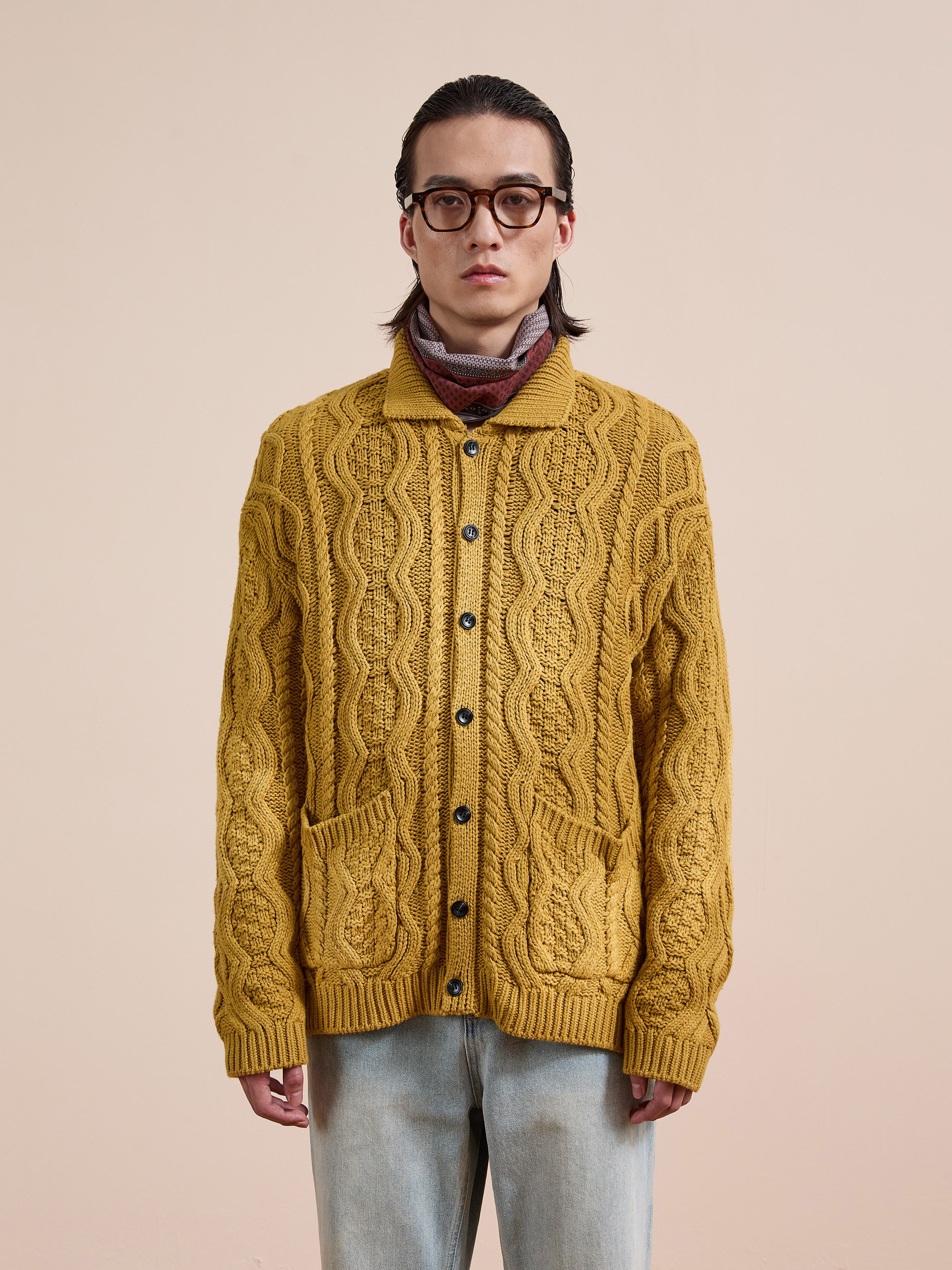 A person with long hair and glasses wears the FOUND Cable Knit Buttoned Cardigan in mustard yellow, paired with a striped scarf and light blue jeans, standing against a plain beige background.