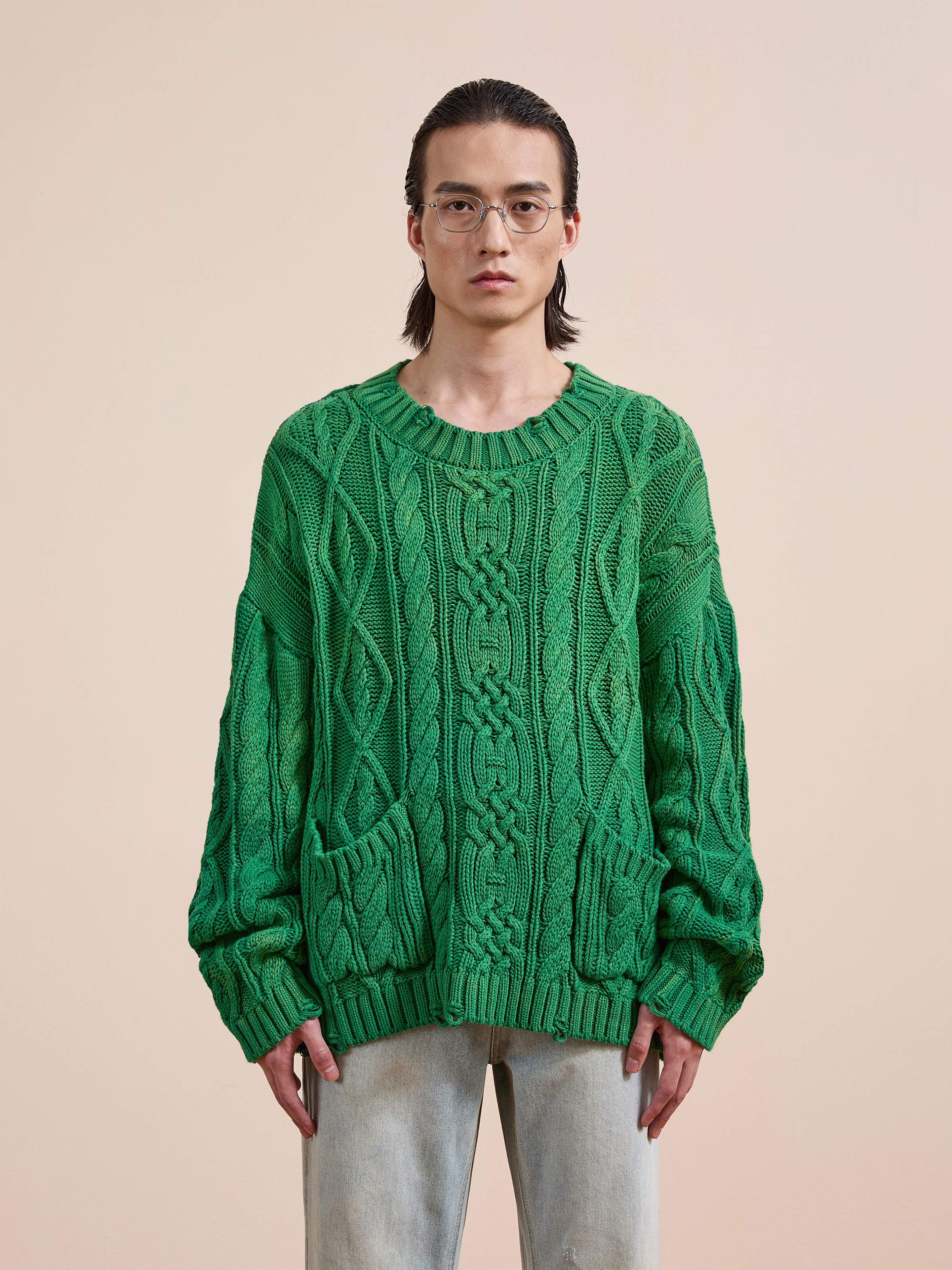 A person with long dark hair and glasses, wearing the Found Cable Knit Pullover Sweater in green with two front pockets and light jeans, stands against a plain beige background.