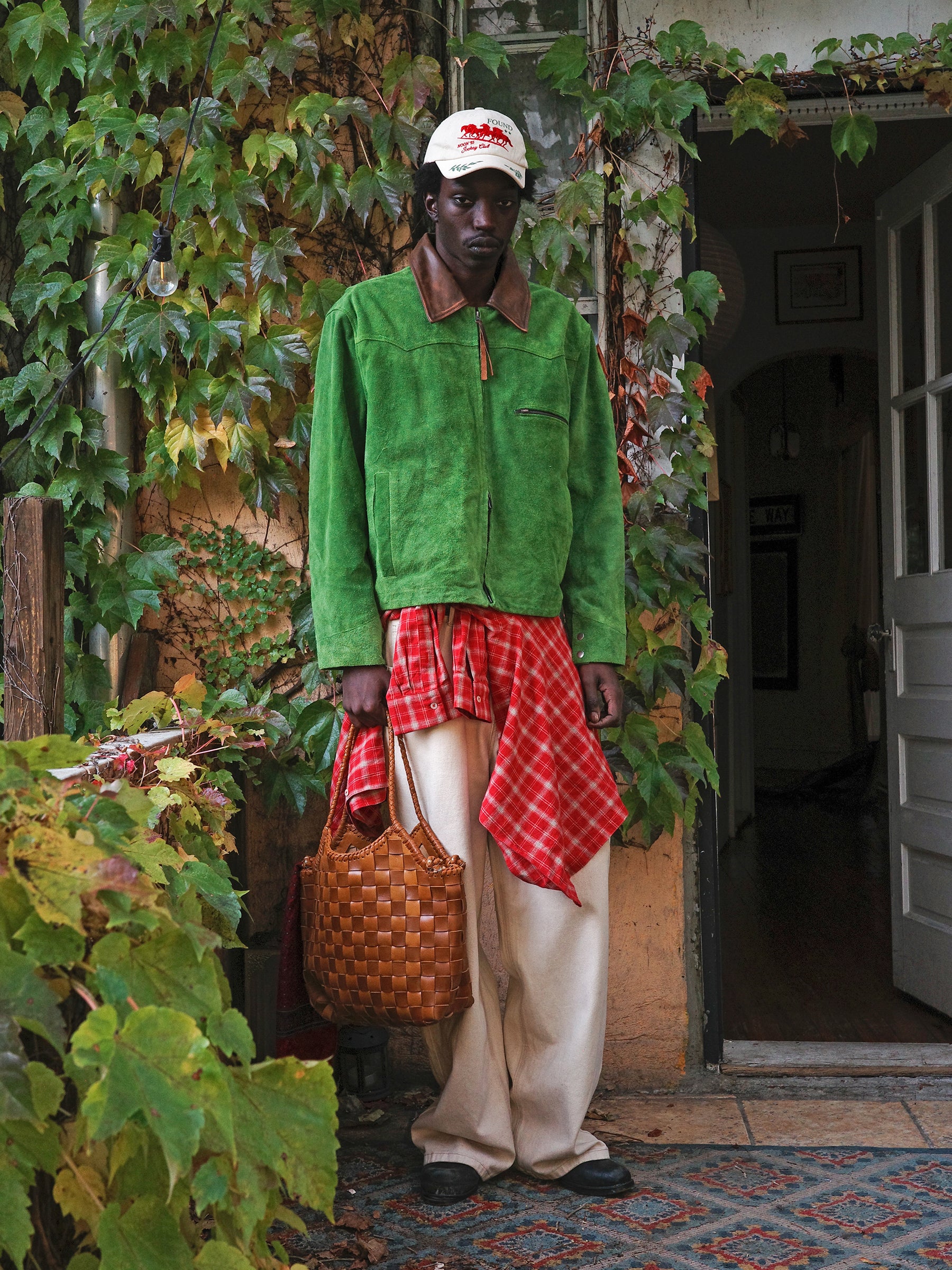 A person stands before a vine-covered doorway, wearing the FOUND Suede Western Barn Jacket, beige pants, a red plaid shirt tied at the waist, a white cap, and carrying a woven bag.