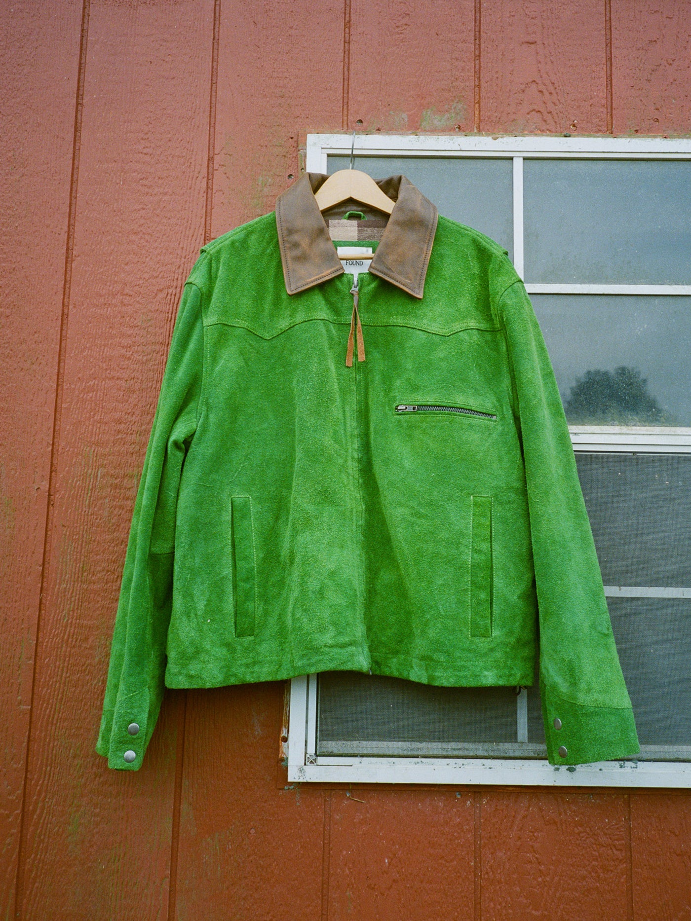 The FOUND Suede Western Barn Jacket, featuring a green suede body, brown collar, and western yoke stitching, hangs on a wooden hanger in front of a red wall and window.