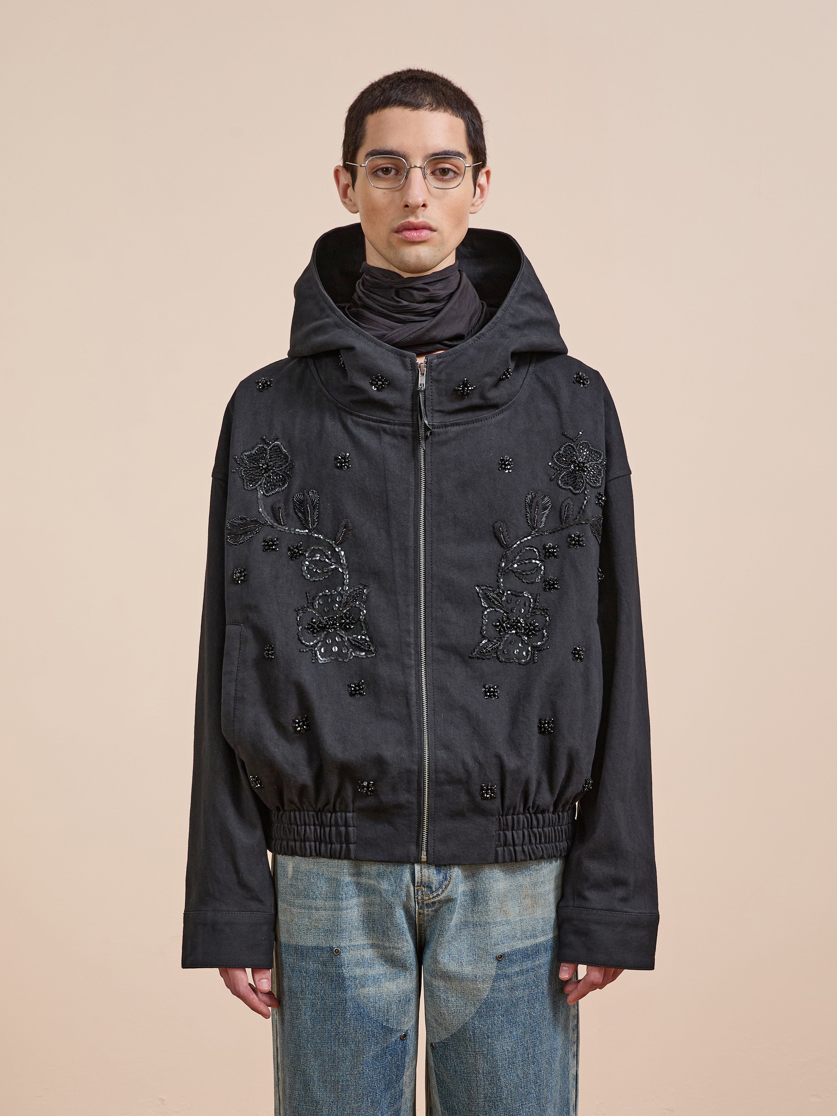 A person wearing glasses, a dark scarf, blue jeans, and the FOUND Hand Embroidered Tonal Anorak Jacket stands against a plain beige background.