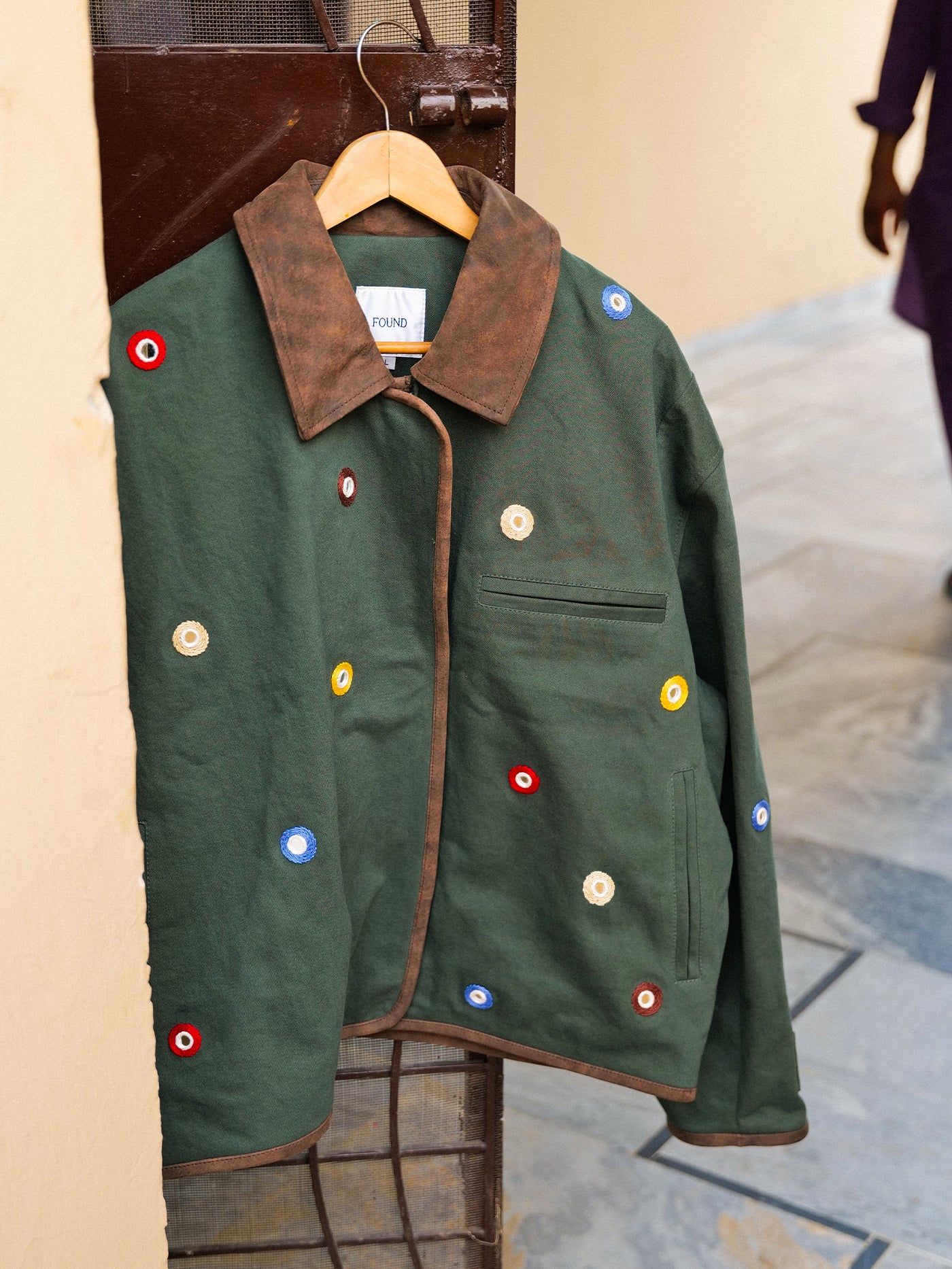 The Found Hand Embroidered Mirror Abhala Jacket, featuring green cotton fabric, a brown collar, and vibrant circular patches, hangs on a wooden hanger by a metal gate near a beige wall.