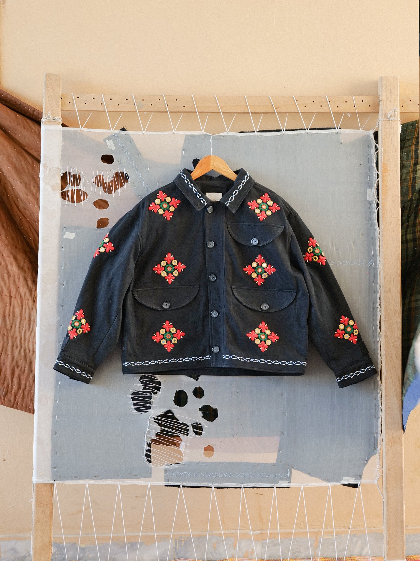 The Found Mirror Ornament Hand Embroidered Work Jacket is showcased on a hanger against a light gray, patchwork textile backdrop, featuring intricate red and green floral embroidery on handcrafted cotton twill.