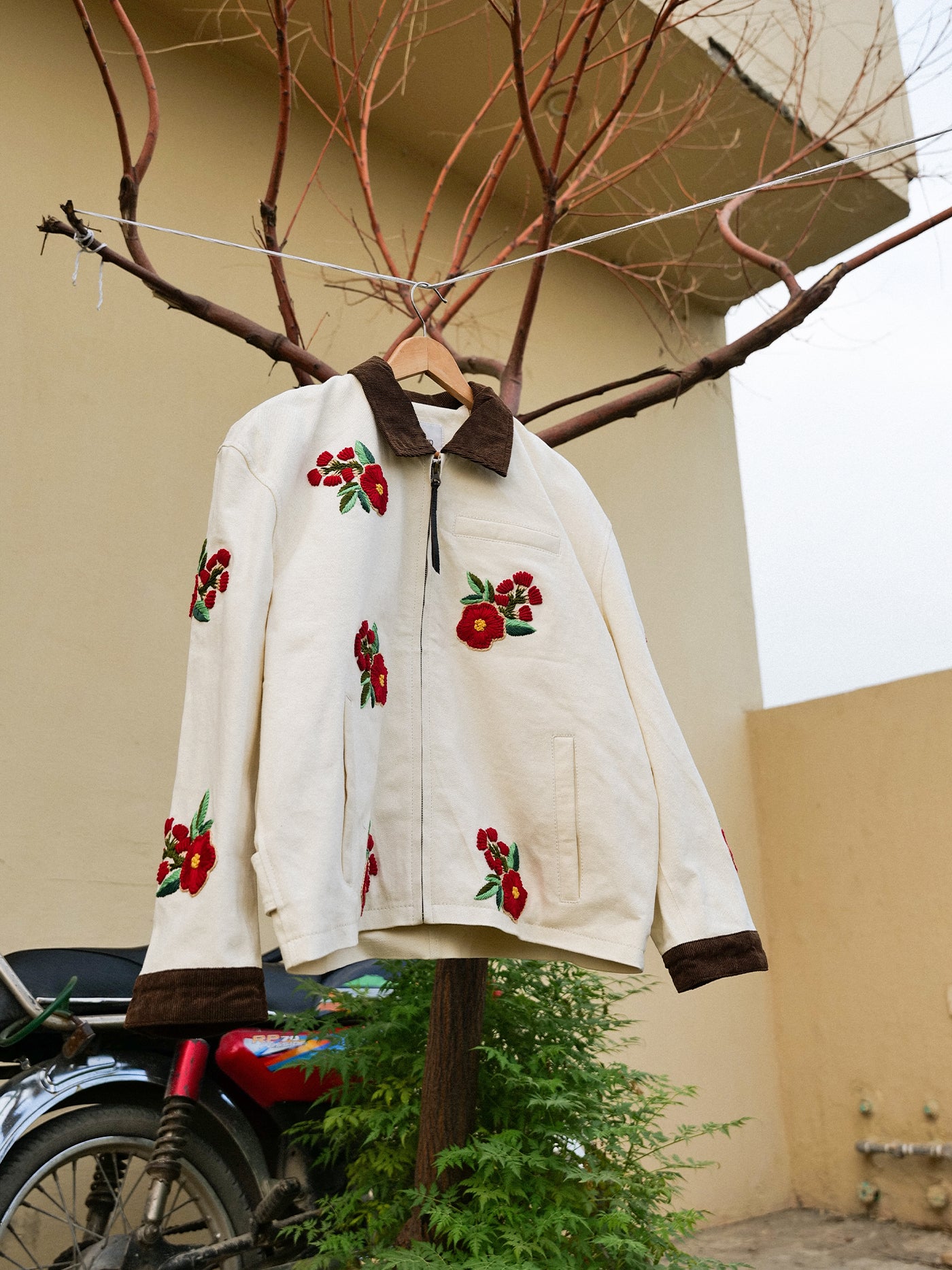 The Floral Hand Embroidered Work Jacket by Found, featuring a cream base with brown collar and cuffs, hangs outdoors from a tree branch, displaying intricate South Asian needlework with a motorcycle and beige building behind.