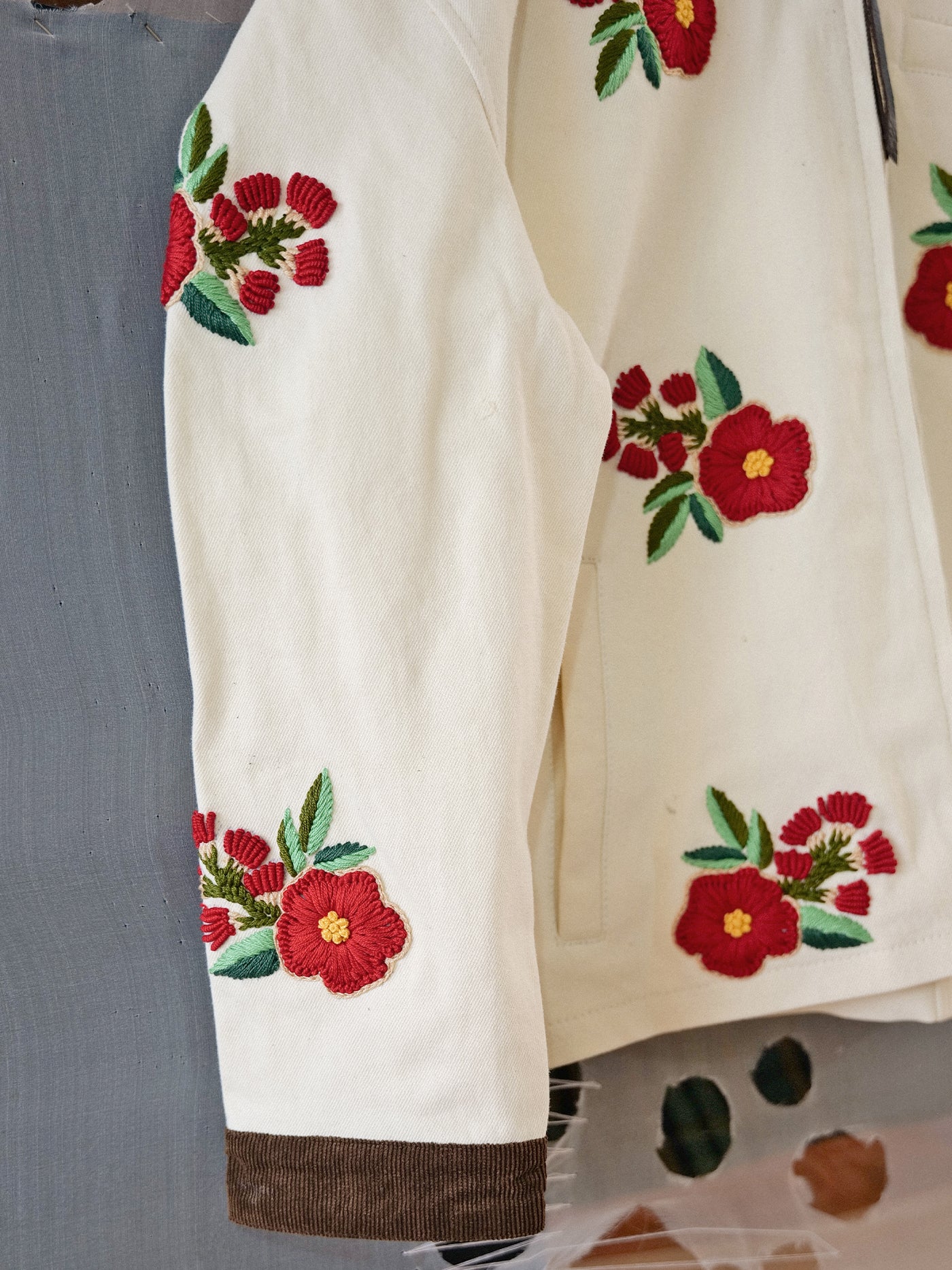 The Floral Hand Embroidered Work Jacket by Found features cream fabric with red and yellow South Asian embroidery on the sleeves and shoulder, green leaves, and a brown cuff.