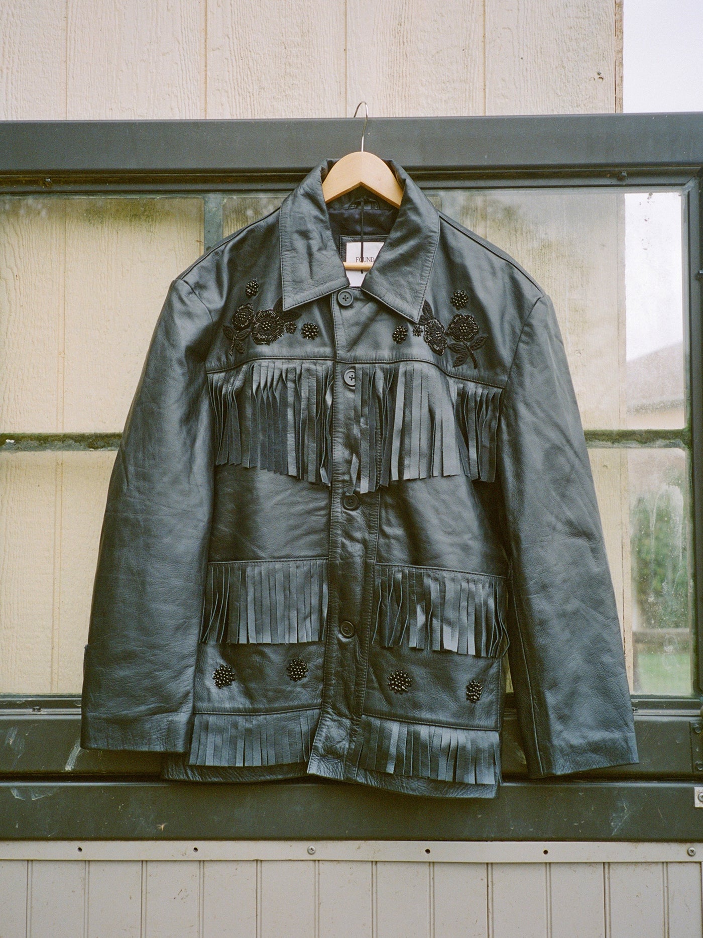 The FOUND Hand Embroidered Leather Fringe Jacket, crafted from genuine cowhide and detailed with floral embroidery, hangs on a wooden hanger in front of a window.