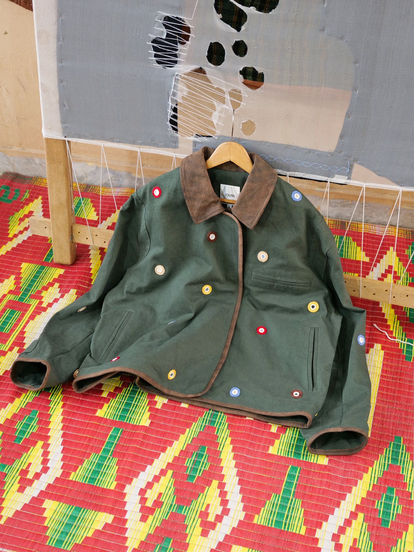 The Found Hand Embroidered Mirror Abhala Jacket is shown on a wooden hanger with a red patterned mat beneath it, set against a distressed fabric backdrop.