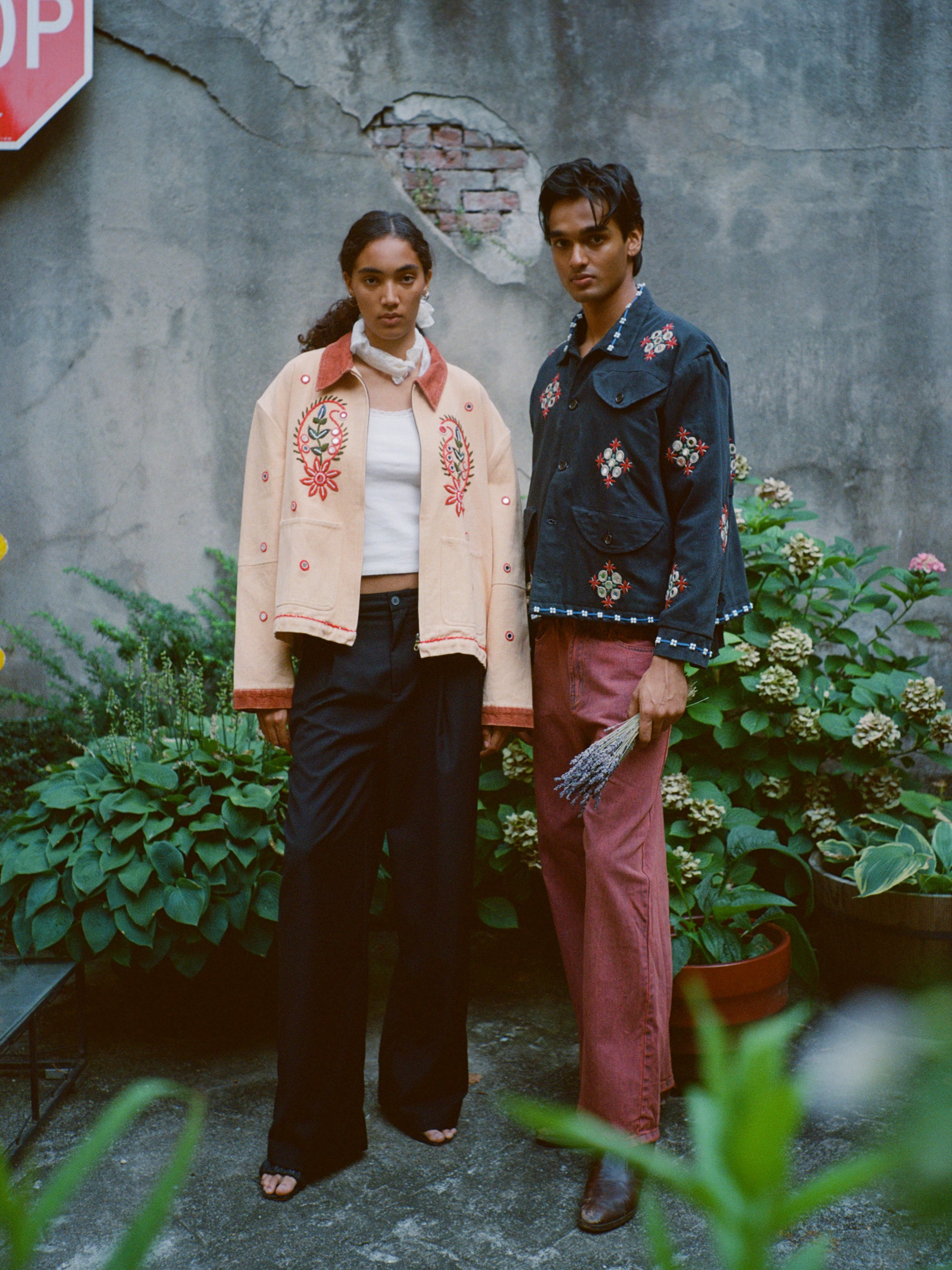 Two people stand by a weathered wall in a garden, both wearing Found’s Paisley Motif Hand Embroidered Work Jacket with wide-legged pants. One holds lavender, surrounded by potted plants and a stop sign in the background.