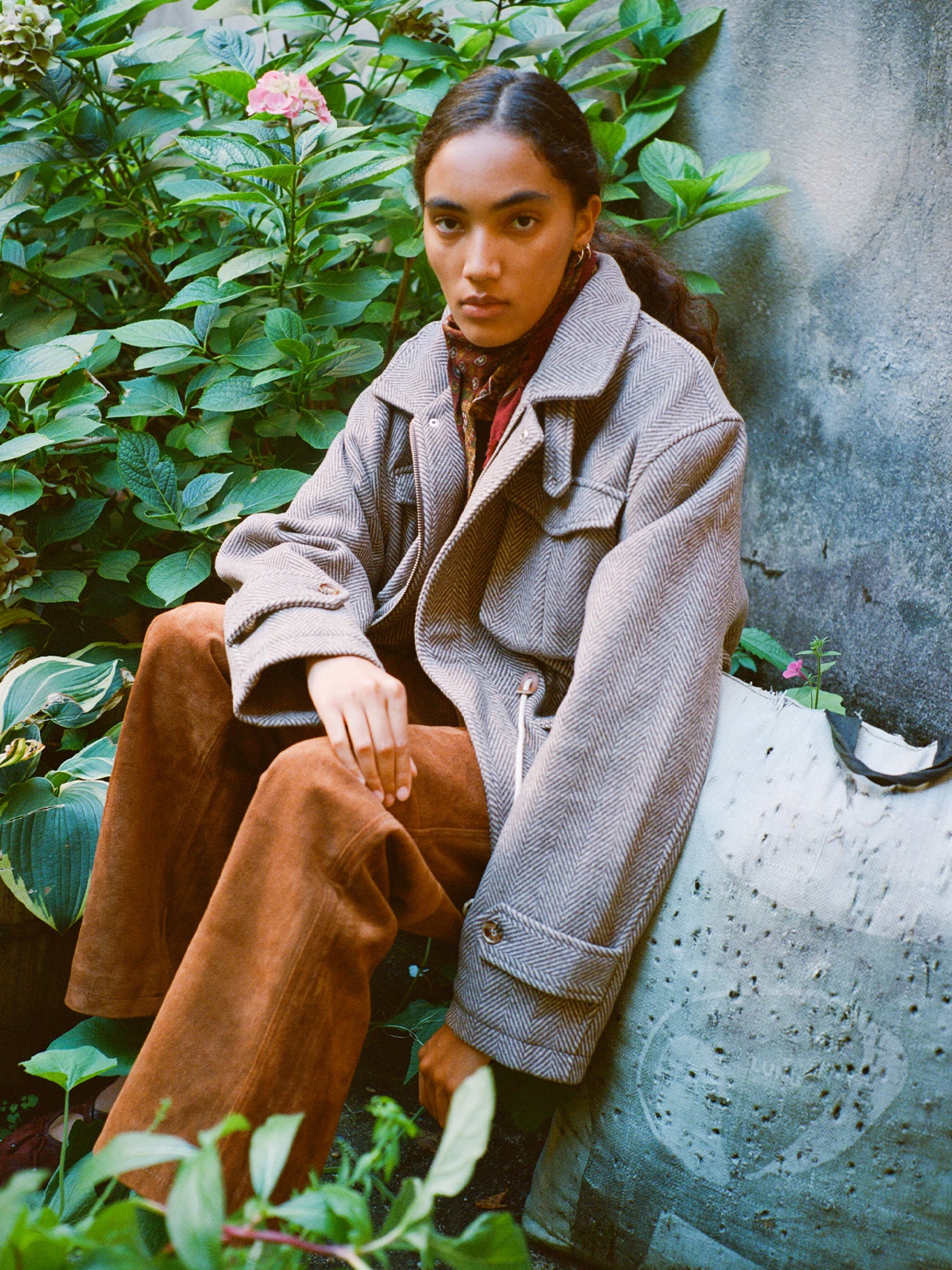 A person with long dark hair sits outdoors among green plants, wearing FOUND’s Herringbone Wool Barn Jacket (unisex relaxed fit), brown pants, and a scarf, looking directly at the camera.