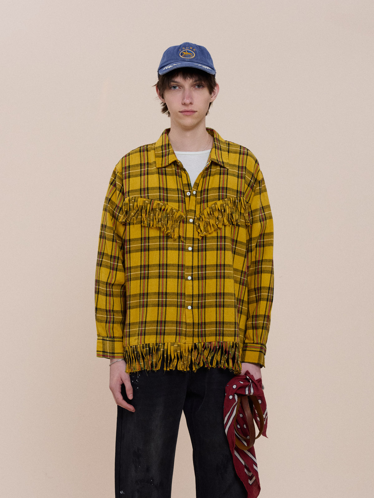 Person wearing the FOUND High Plains Fringe Shirt in yellow plaid, black pants, a blue cap, and holding a red bandana against a beige background.