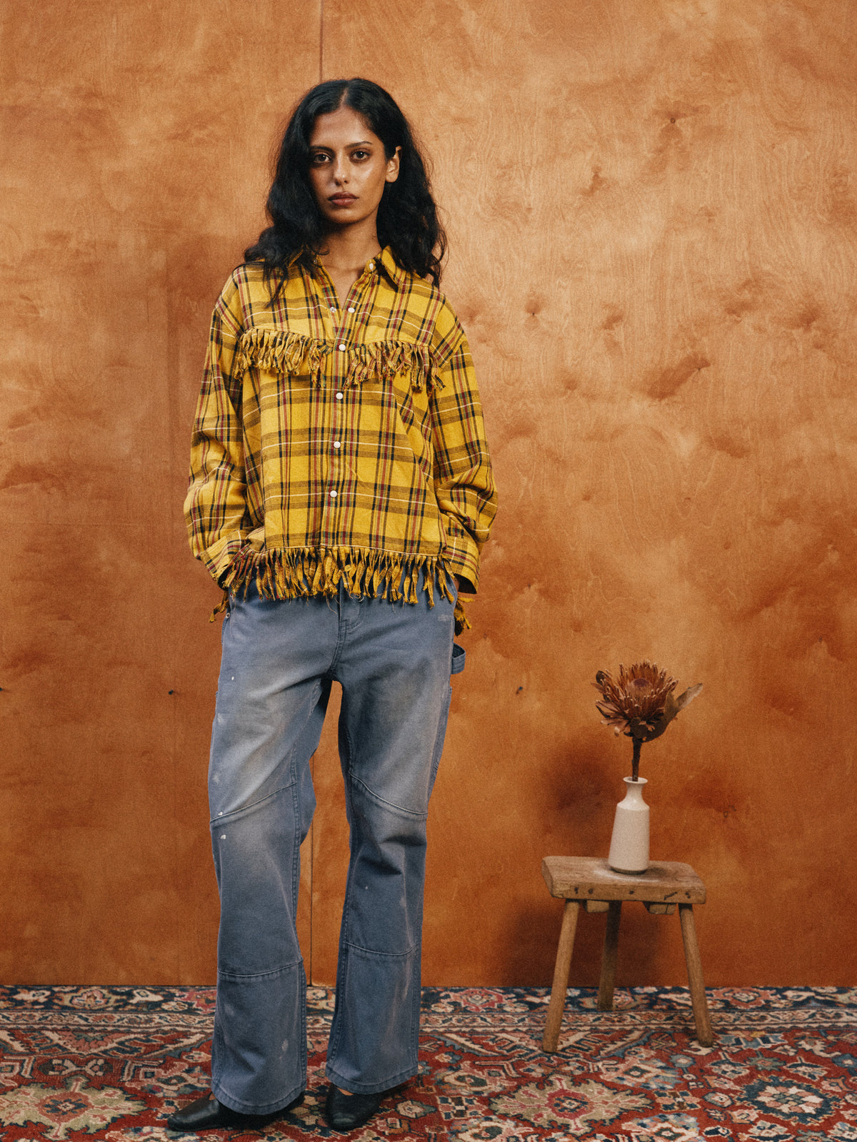 A person stands indoors by a wood-paneled wall, wearing the FOUND High Plains Fringe Shirt and loose blue jeans. A stool with a vase and dried plant is next to them.