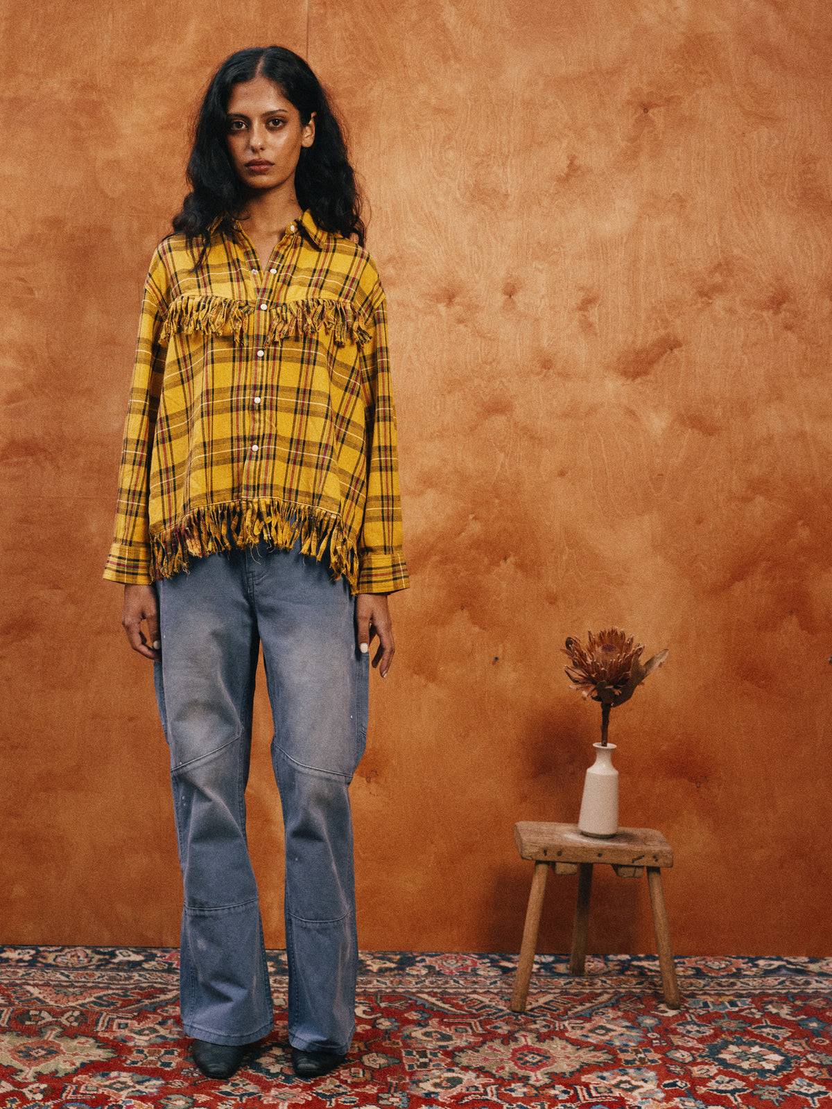 A person stands indoors in the FOUND High Plains Fringe Shirt and blue jeans beside a stool with dried flowers, set against an orange-brown wall.