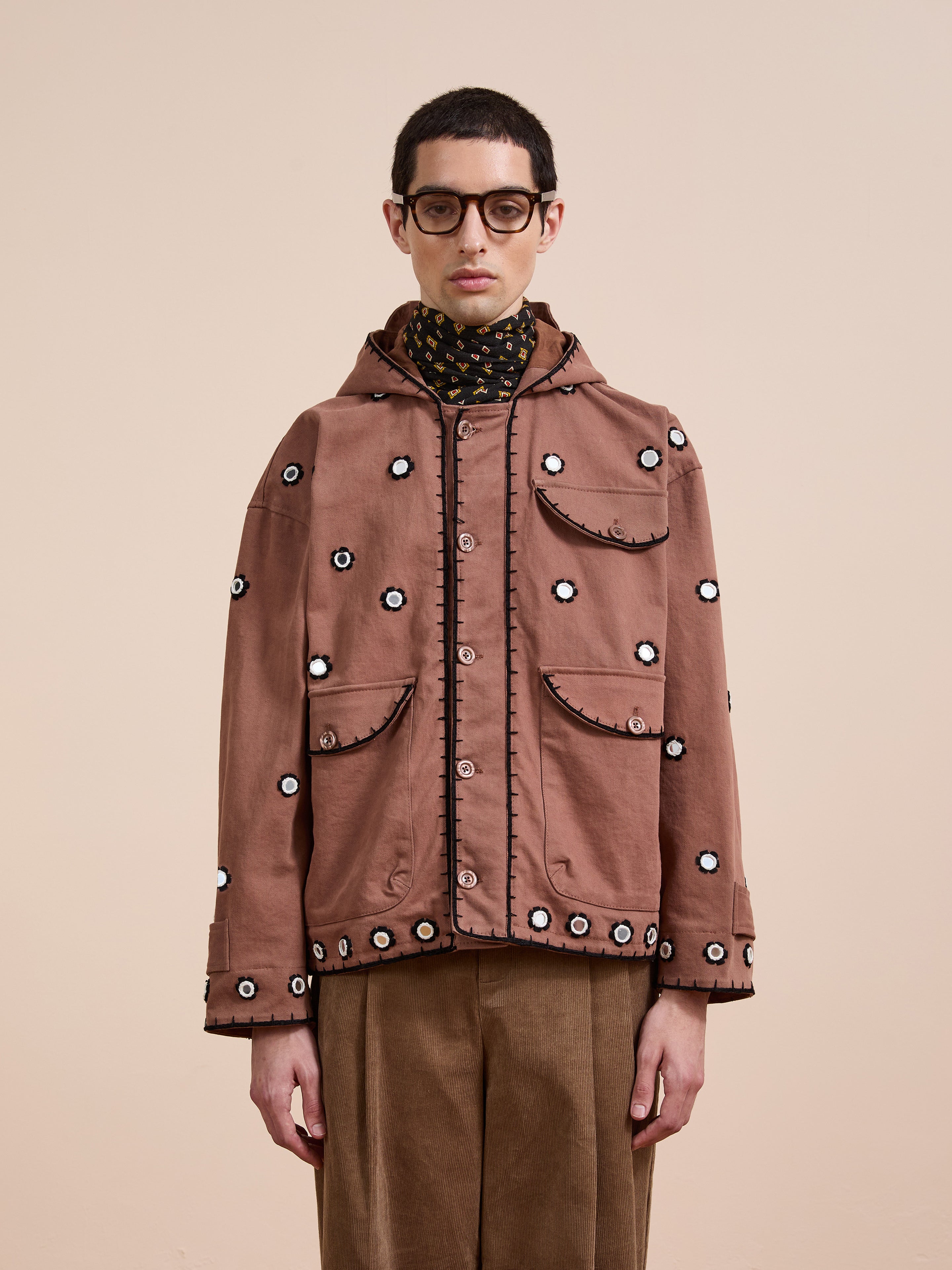 A person wearing glasses, the FOUND Hand Embroidered Hooded Mirror Anorak, a black patterned scarf, and brown pants stands against a beige background.