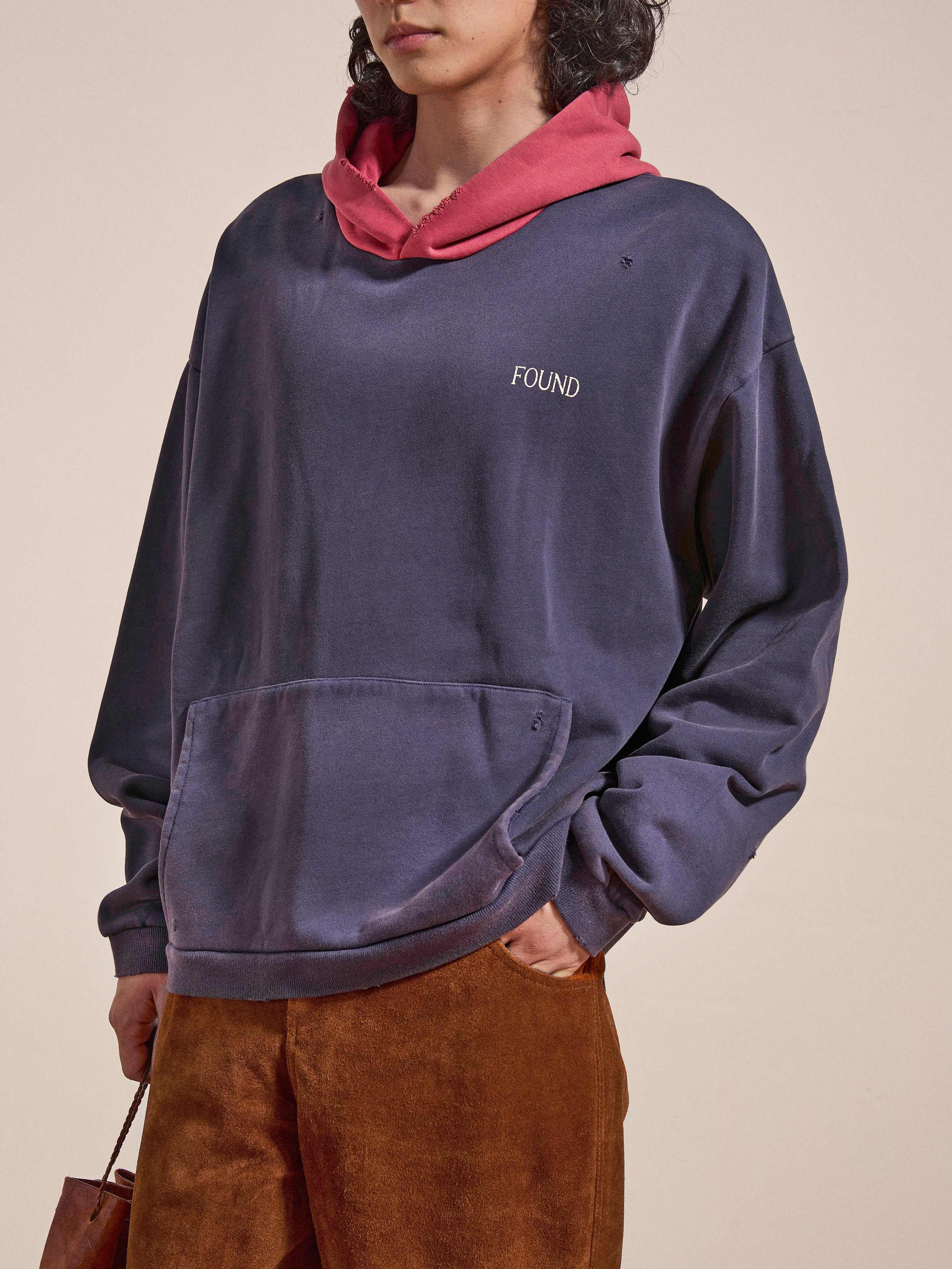 Individual wearing FOUND’s Distressed Logo Hoodie in navy blue, featuring a pink hood and "FOUND" text, paired with brown pants. The vintage feel of the french terry cotton gives it a relaxed vibe. They are holding a brown bag.