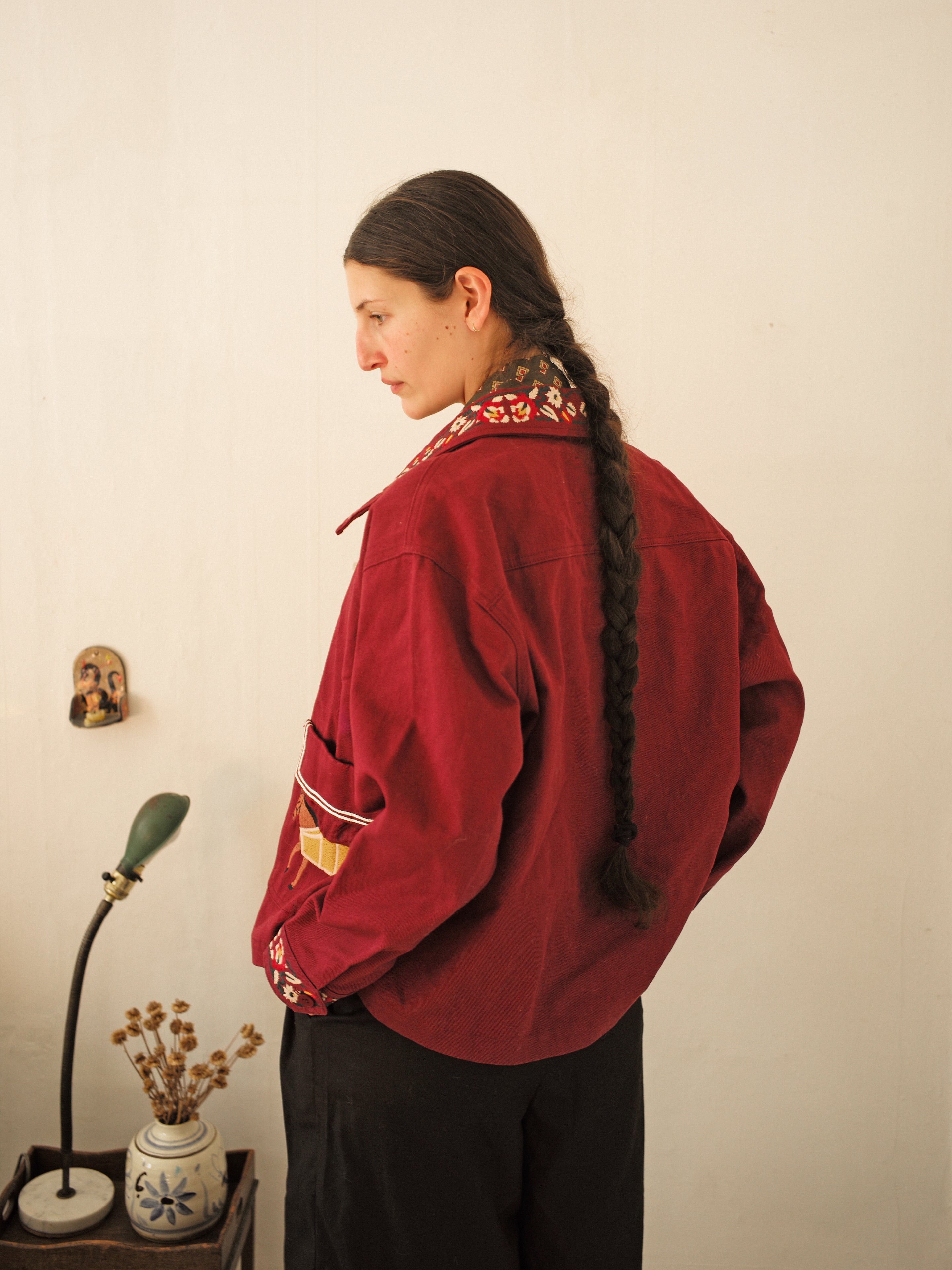 A person with long braided hair stands in an Oxblood Horse Equine Jacket by FOUND, showcasing racing-inspired colors and a patterned collar, facing sideways. A small art piece and a green lamp are seen in the background.
