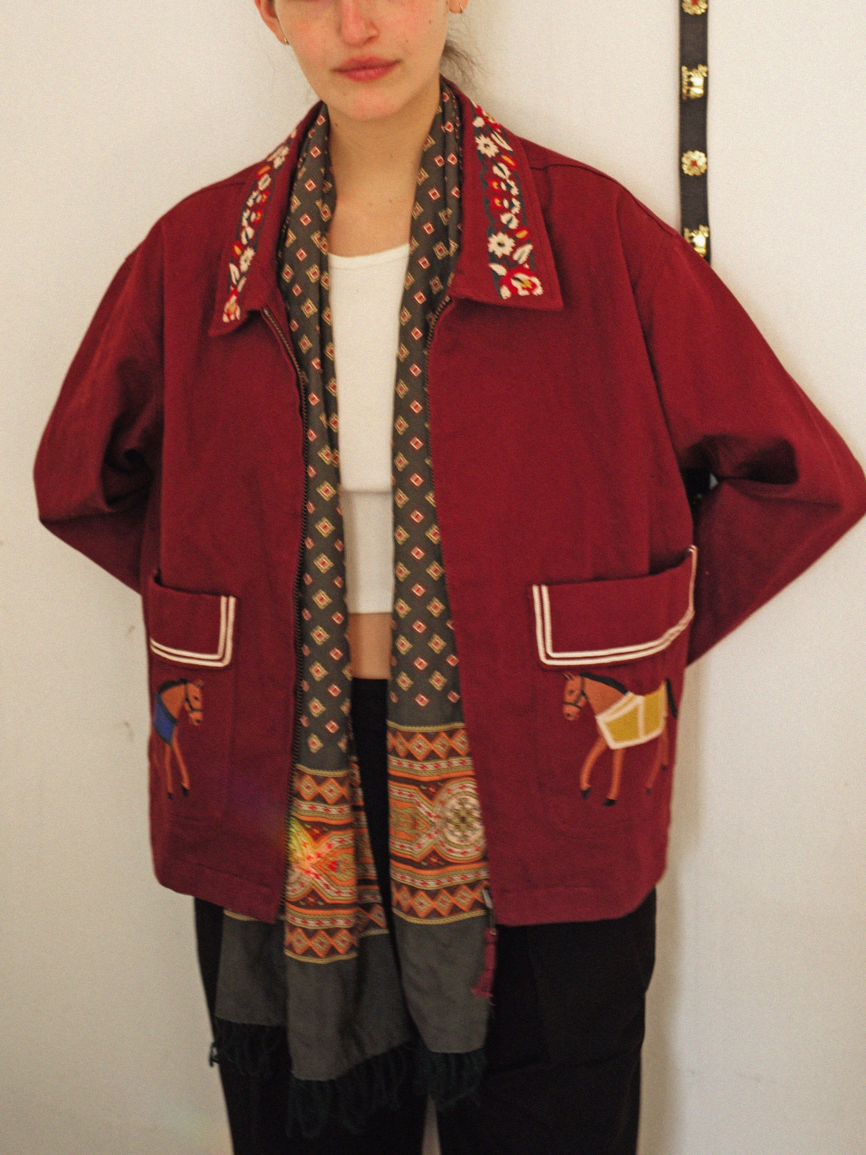 A person wears the FOUND Oxblood Horse Equine Jacket, featuring decorative embroidery and horse motifs inspired by 70's jockey uniforms. It’s styled with a patterned scarf and white shirt, echoing equine work jackets in rich oxblood tones.