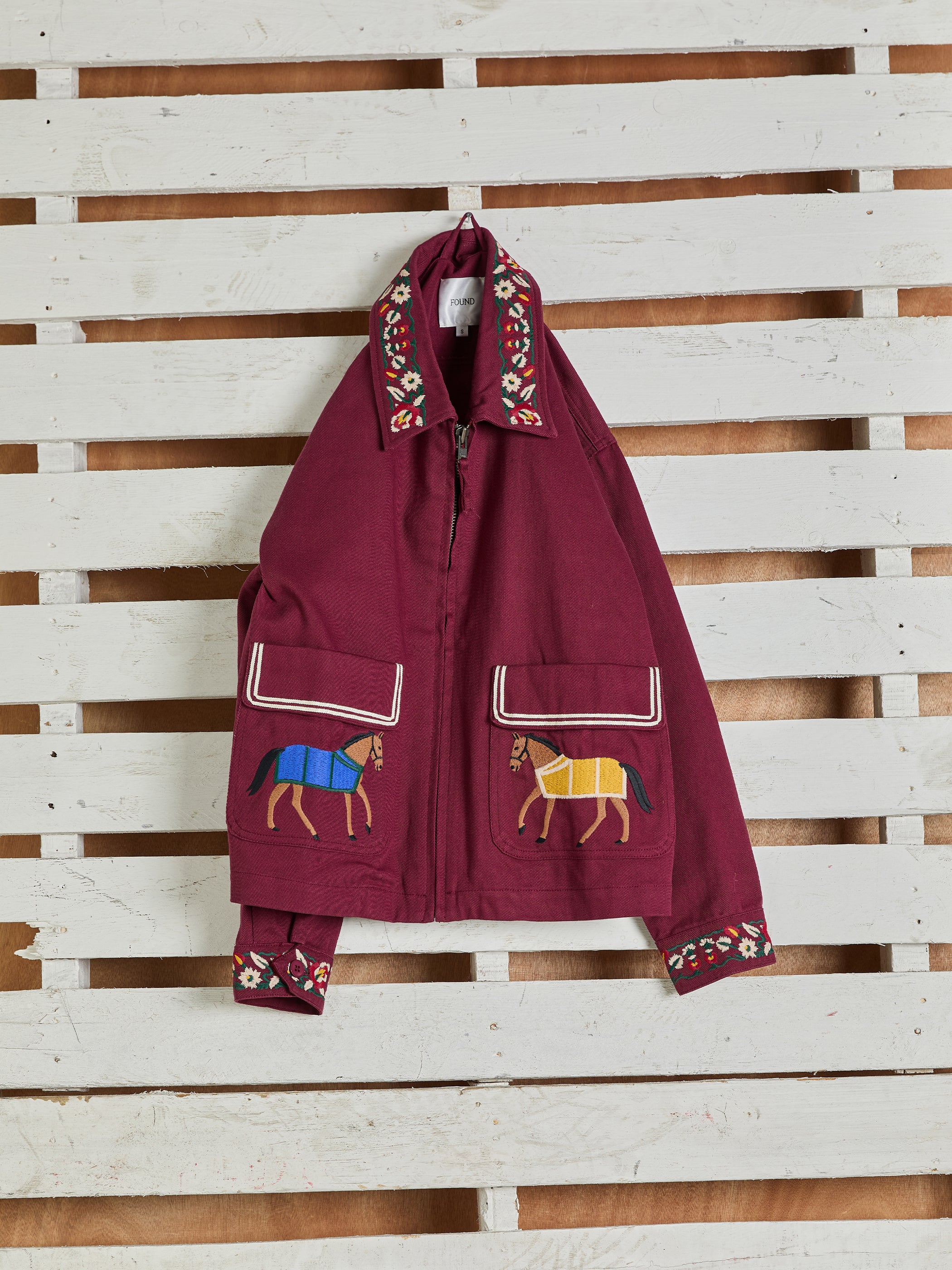 Oxblood Horse Equine Jacket by FOUND, featuring embroidered horses on the front pockets, displayed on a white wooden wall.