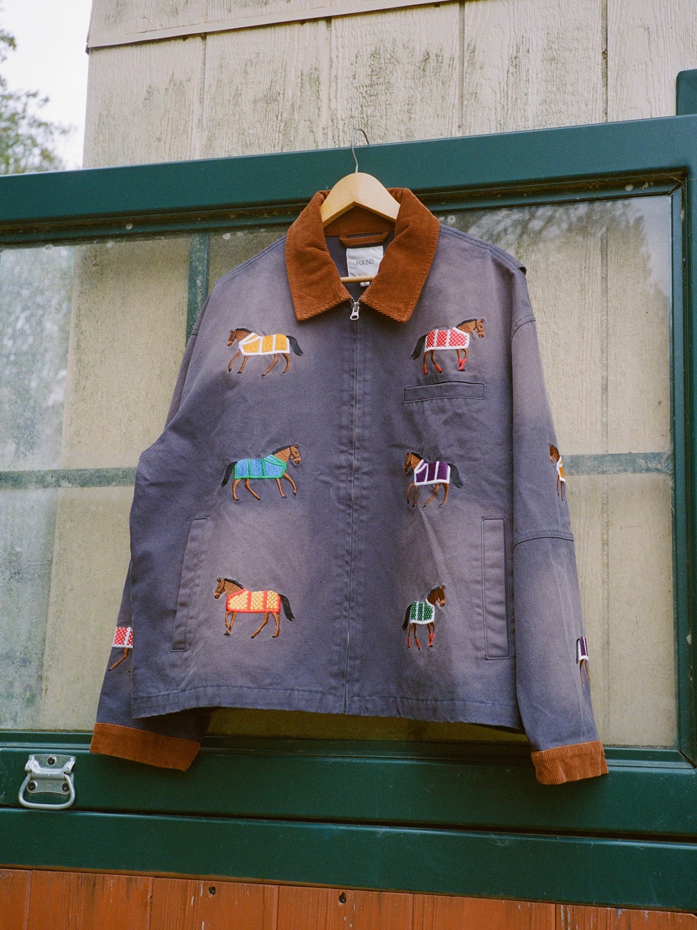 The Found Lariat Horse Embroidered Work Jacket, a unisex navy piece with a brown collar and cuffs and colorful horse embroidery, hangs on a wooden hanger in front of a green-framed window.