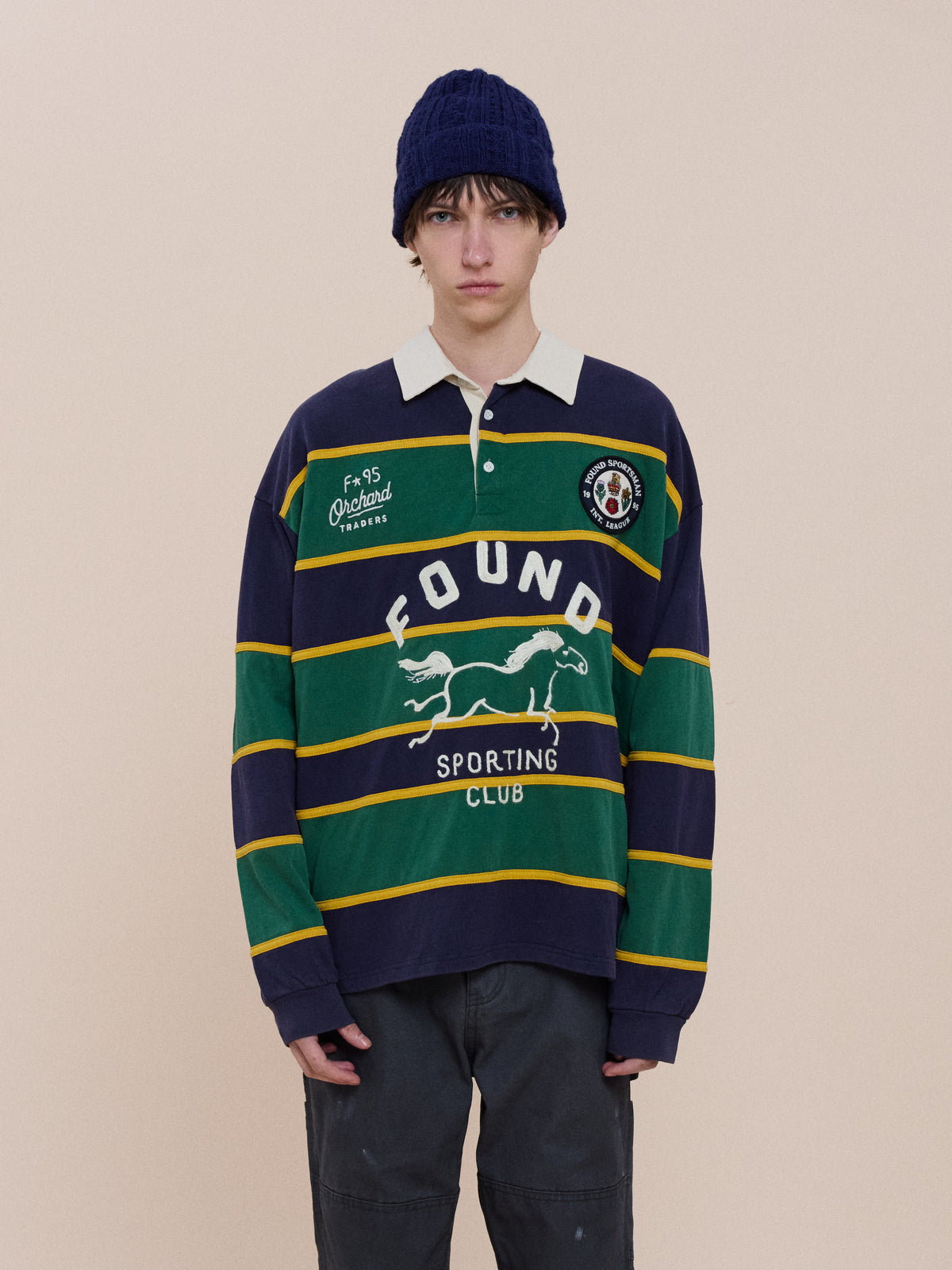 A person in a navy knit beanie and a FOUND Horse Logo Rugby stands against a plain beige background.