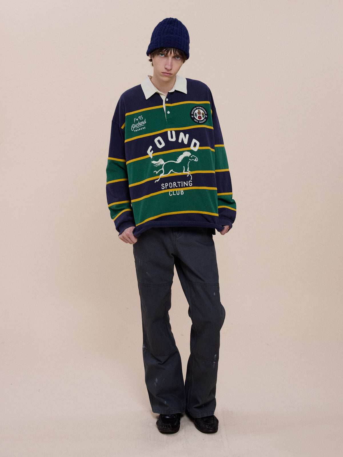 Person wearing a navy beanie, FOUND Horse Logo Rugby, dark pants, and black shoes stands against a plain beige background.