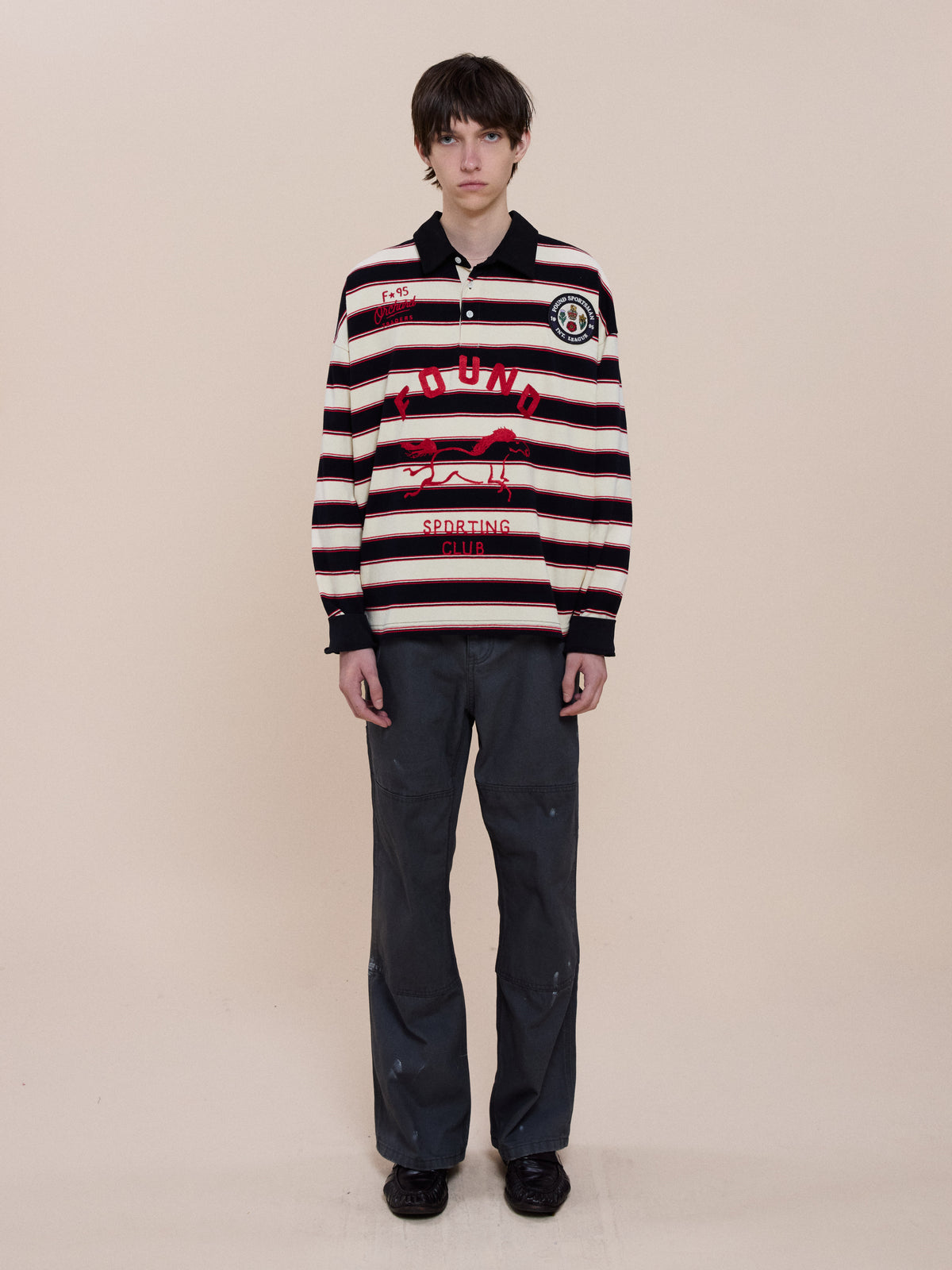 Person stands facing forward in a FOUND Horse Logo Rugby, unisex relaxed fit dark gray pants, and black shoes, against a plain beige background.