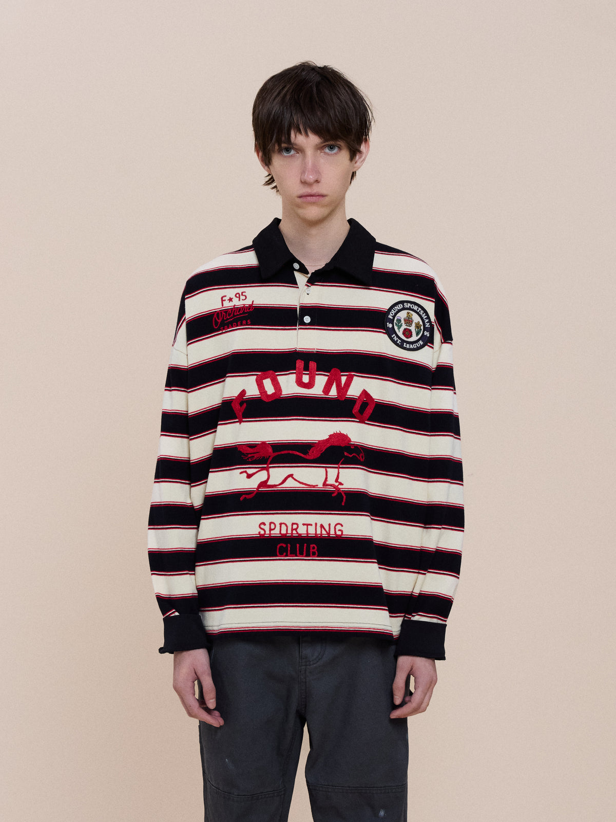 Model wears the FOUND Horse Logo Rugby, a unisex black and white striped shirt with red lettering and graphics, against a plain beige background.