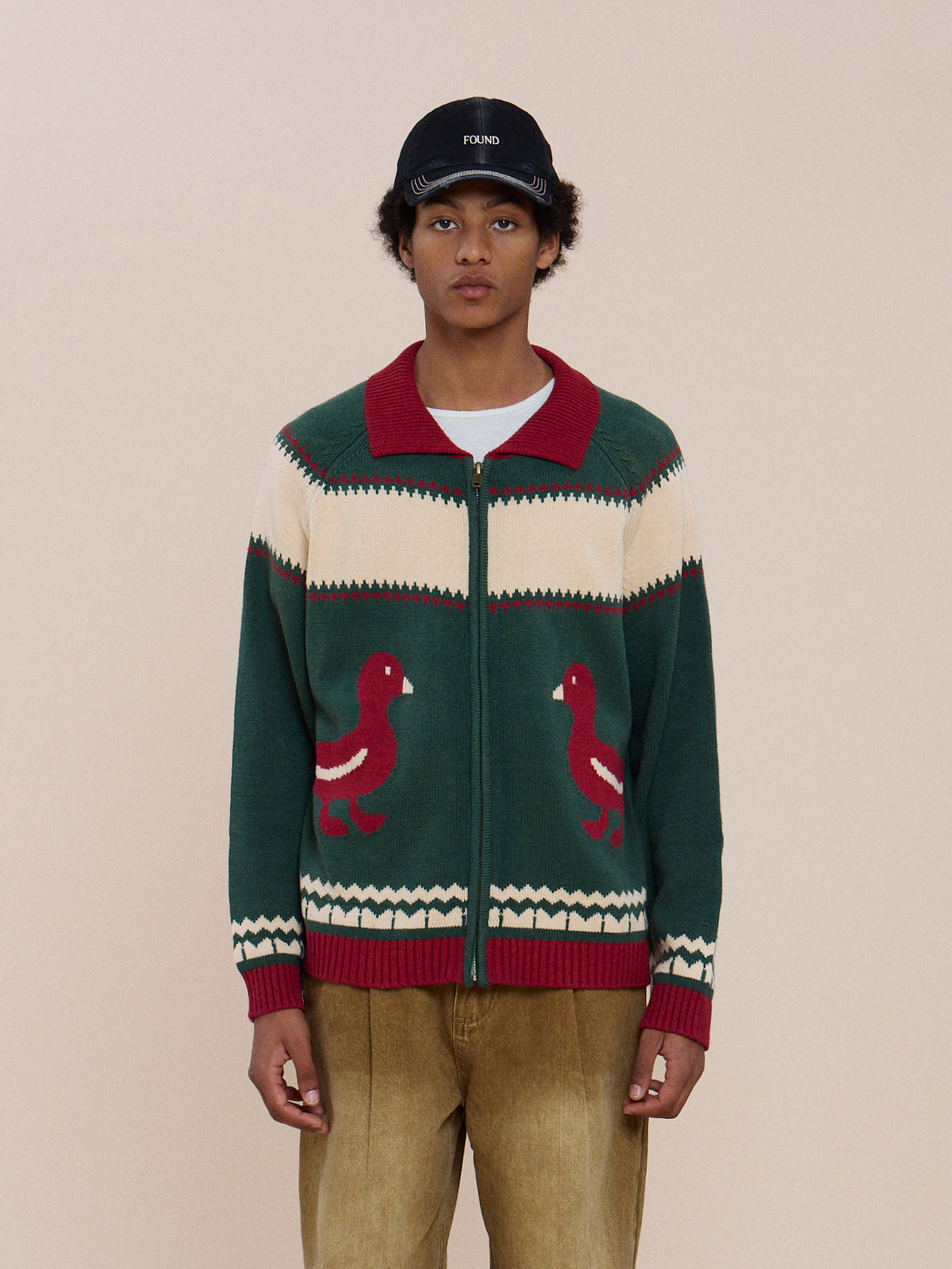 Person in tan pants, FOUND Isles Sweater—unisex vintage knitwear with red and white bird pattern—and a black cap stands against a beige background.